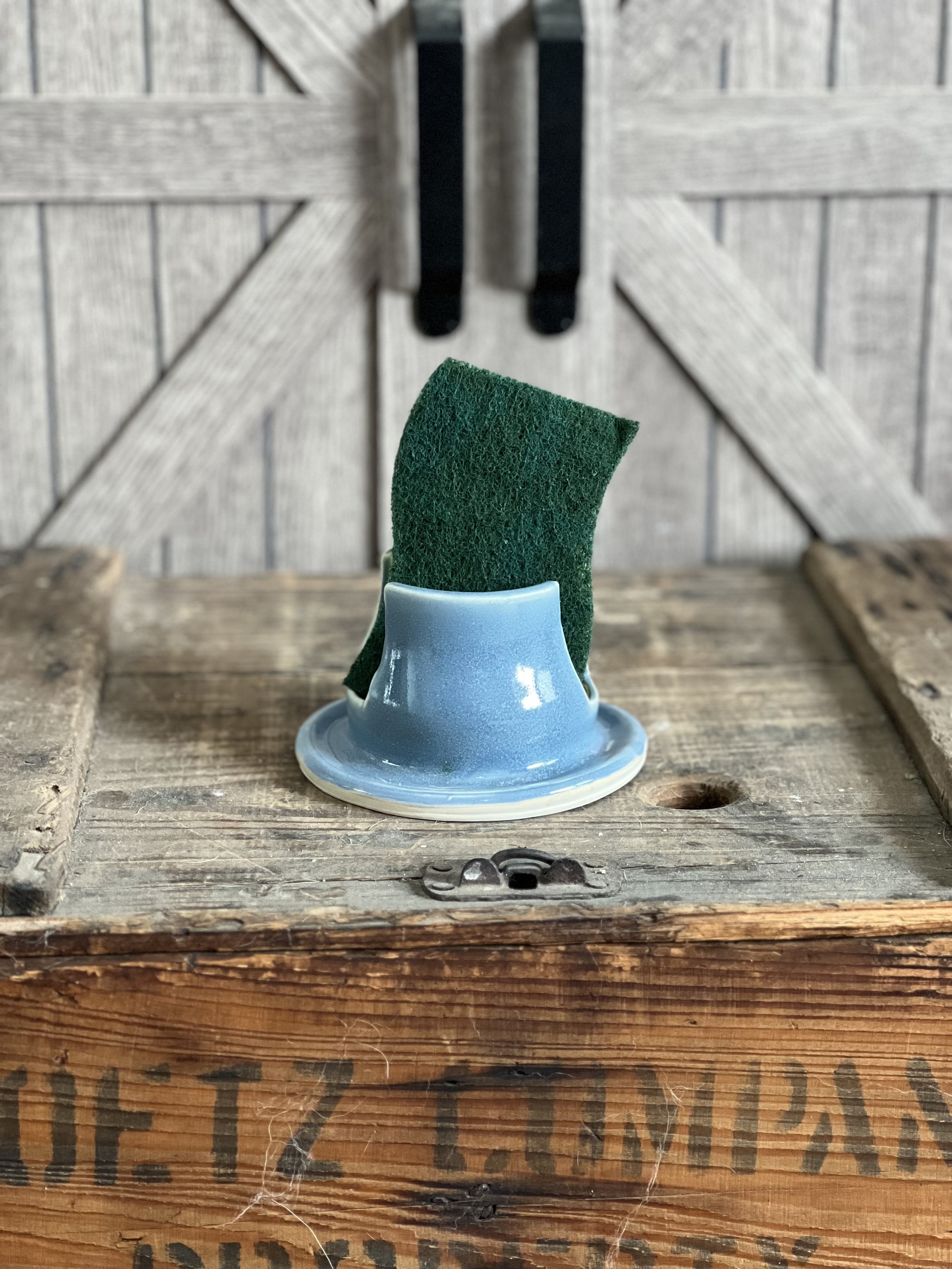 Porcelain cup with a green scrub sponge upside down on top, placed on a rustic wooden table with a background of weathered barn doors.