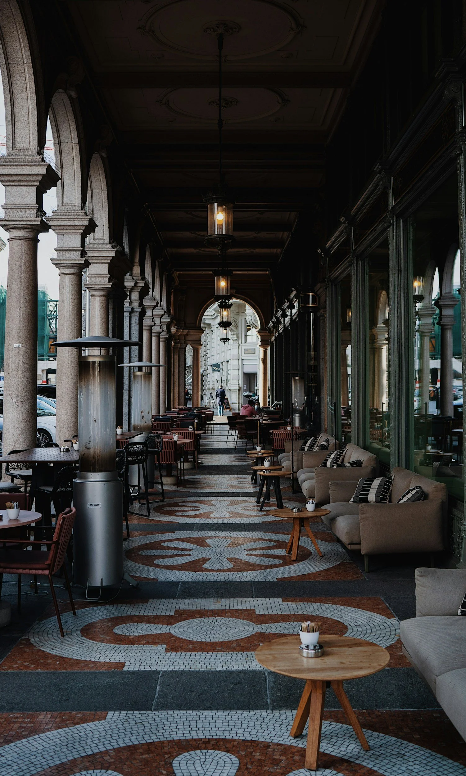 Outdoor cafe with cushioned sofas and small wooden tables under archways with decorative tiled floor