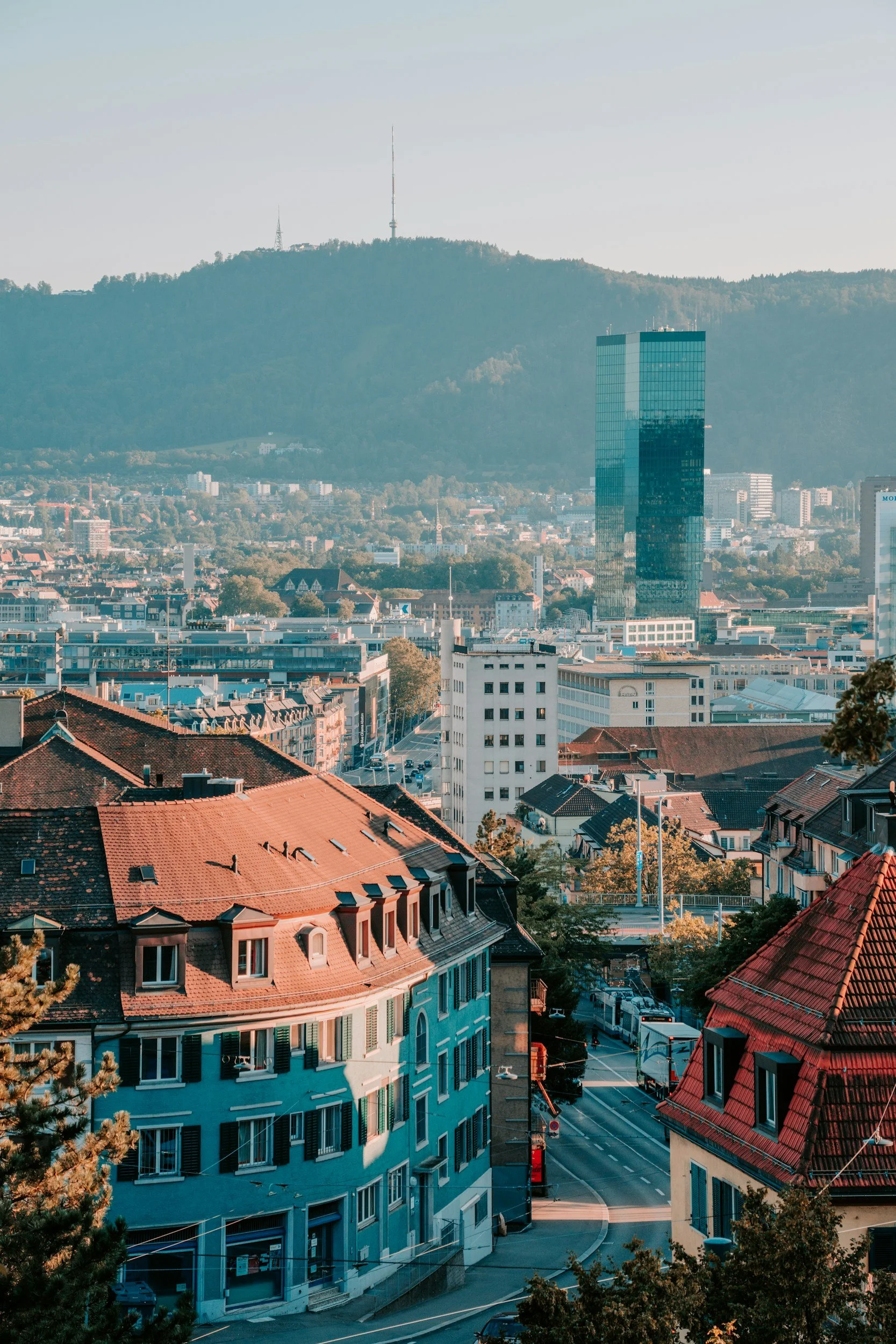 Cityscape with a mix of historic and modern buildings, including a tall glass skyscraper, with hills in the background topped with two radio towers.