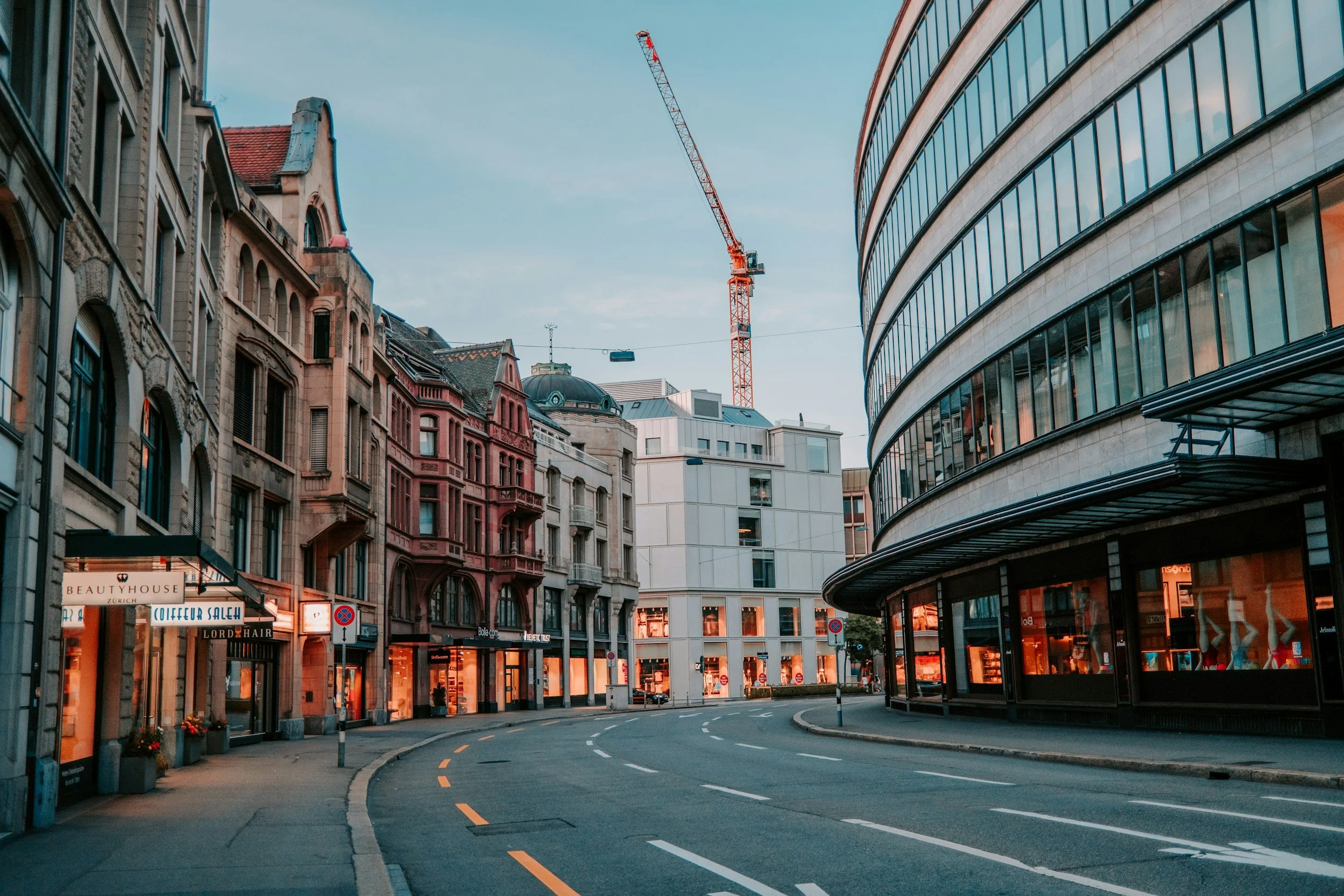 Empty city street lined with shops and historic buildings, with a construction crane in the background, during twilight.