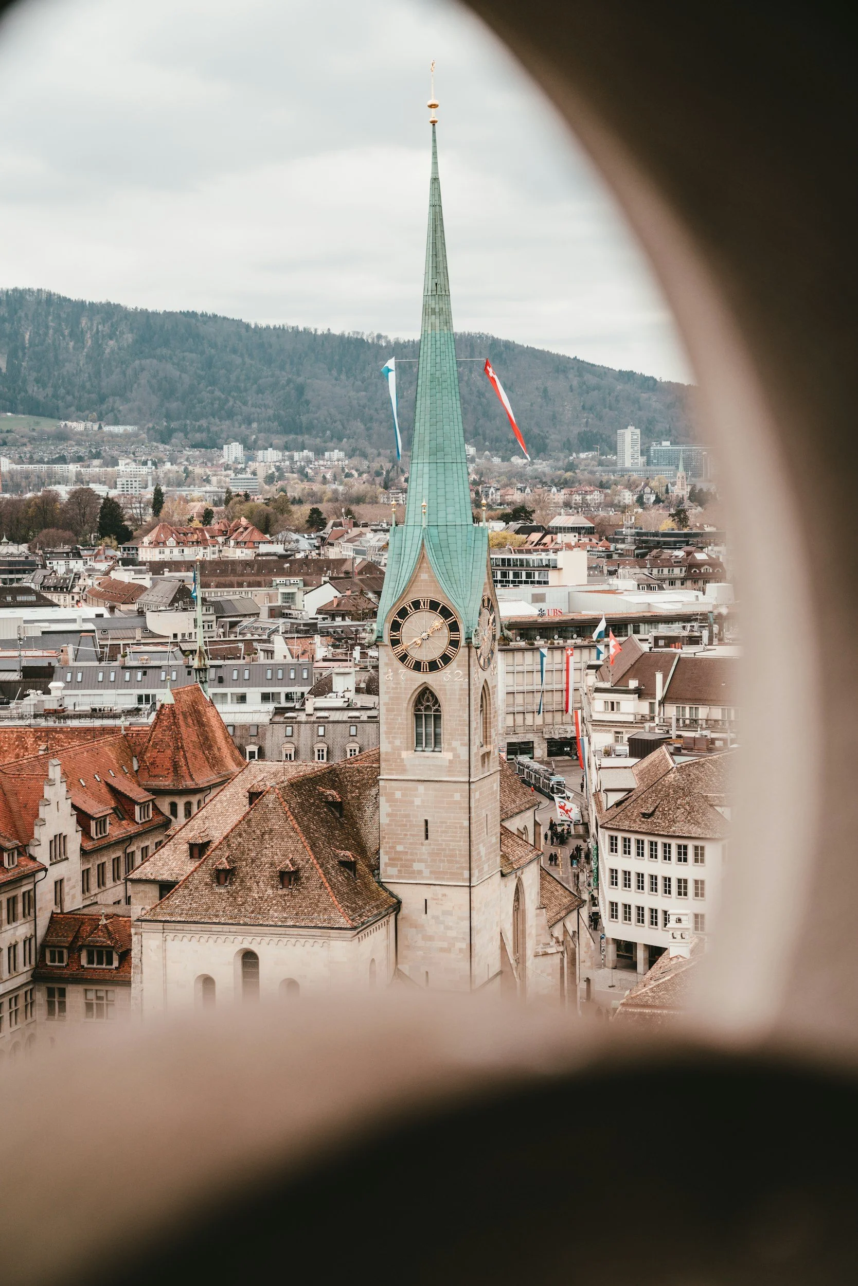 View of a historic clock tower with a green steeple, seen through a small window, with city buildings and mountains in the background.