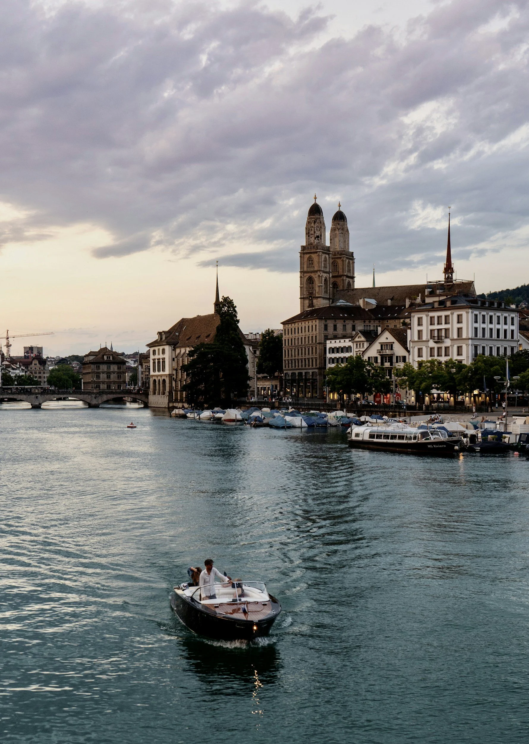 A cityscape with a river, boats, and historic buildings, including a church with twin towers and spires, at sunset or late afternoon.
