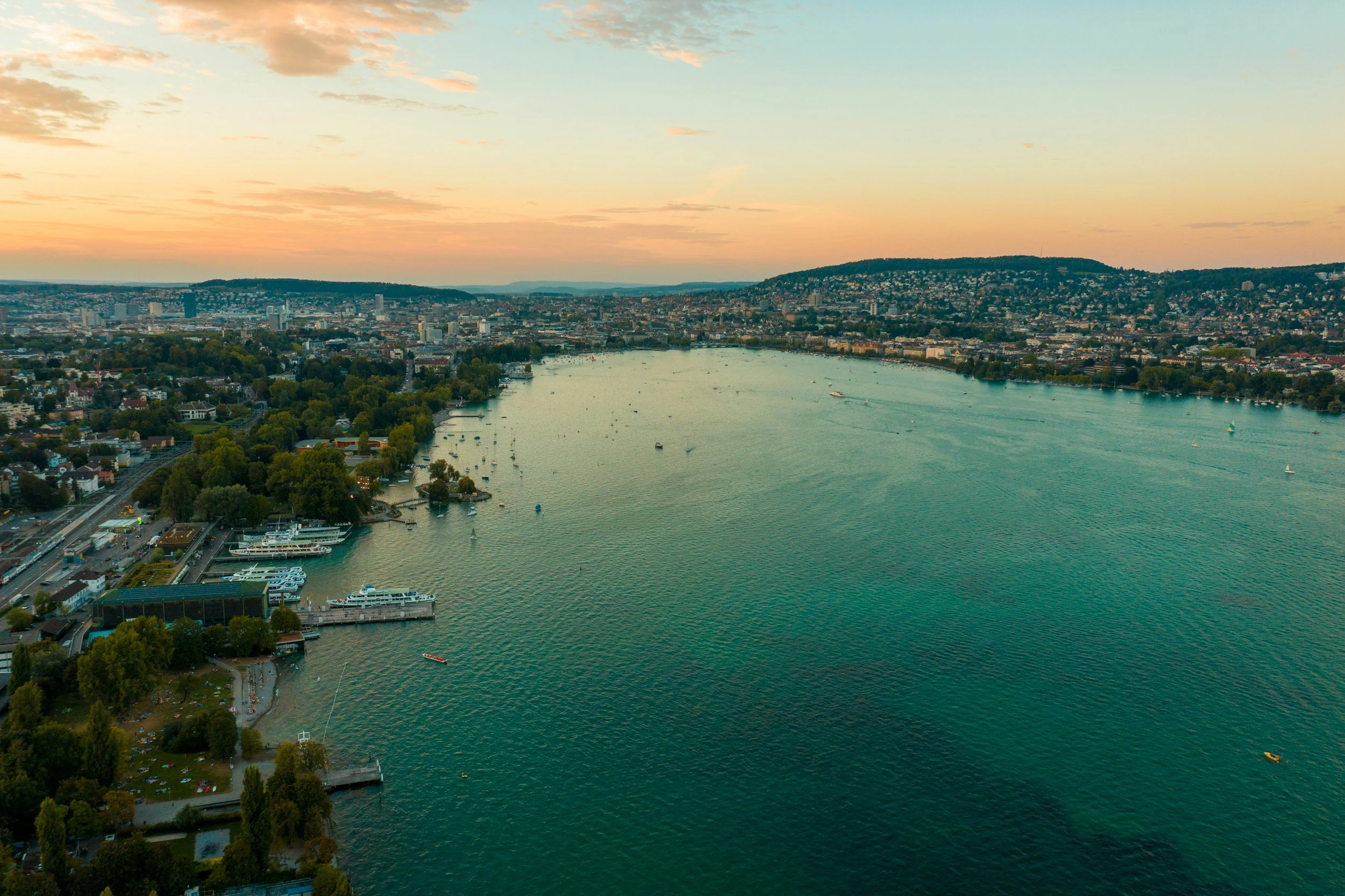 Aerial view of a city with a large river running through it, surrounded by green trees, with boats on the water and a cityscape with buildings and hills in the background at sunset.