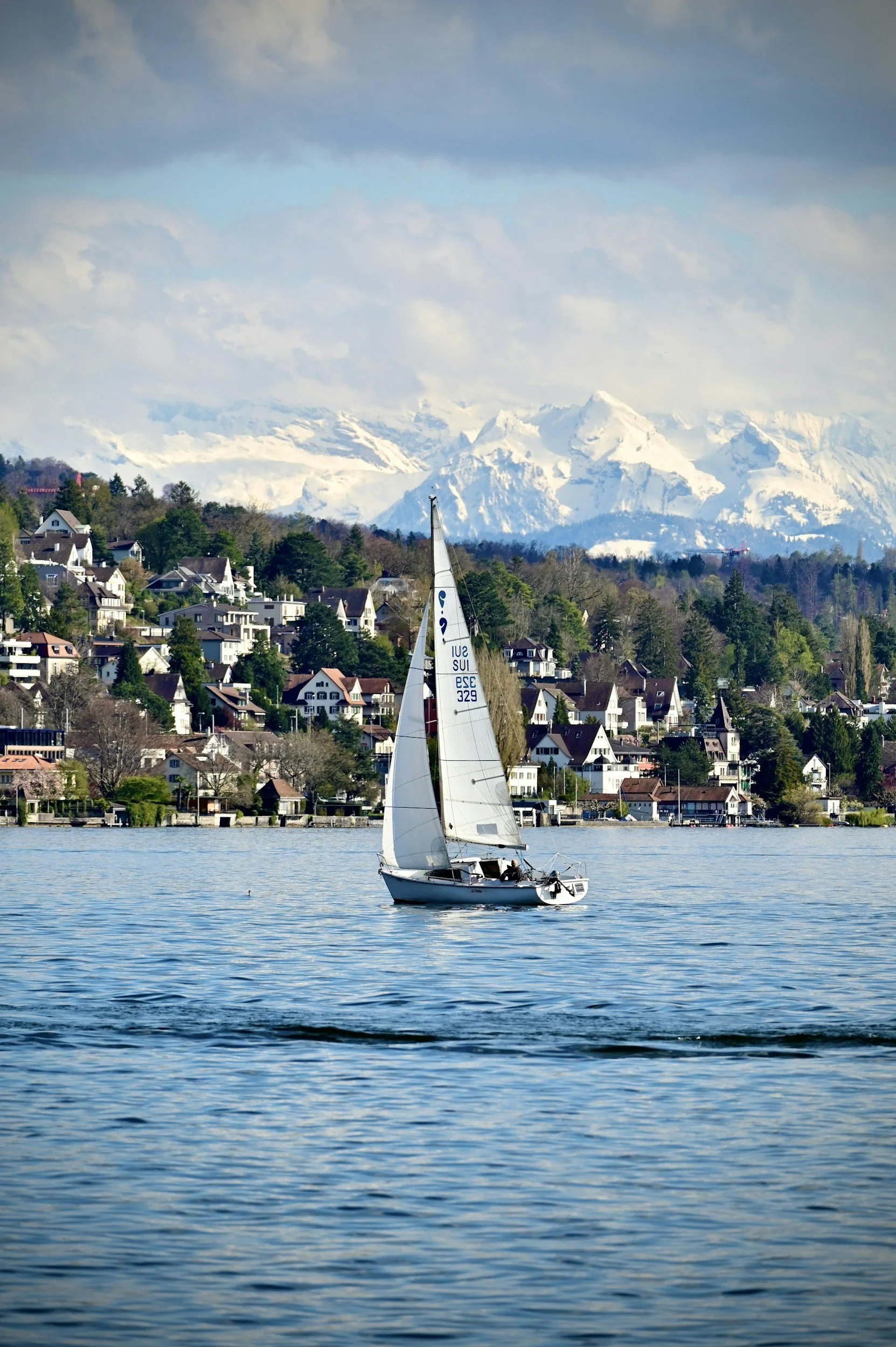 A sailboat on a calm body of water with a town on a hillside and snow-capped mountains in the background.