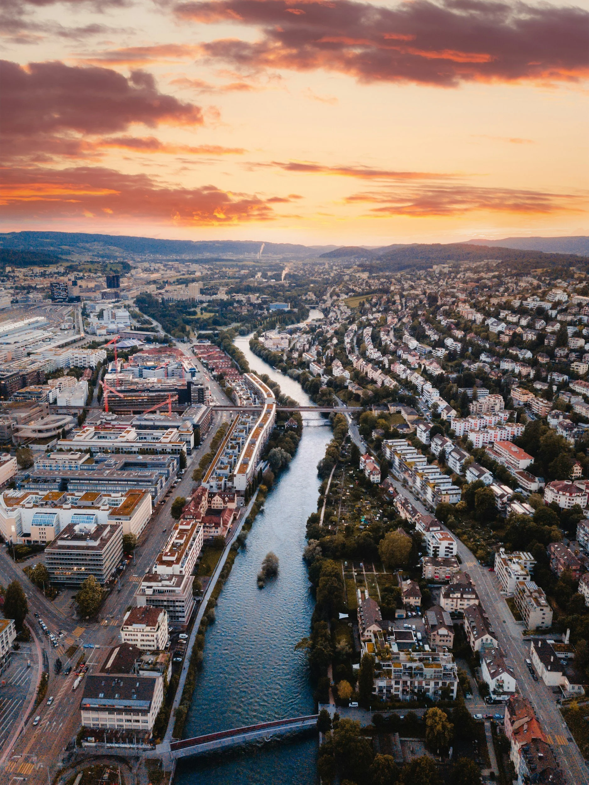 An aerial view of a river flowing through a city at sunset, with buildings, roads, and bridges visible along the riverbanks, surrounded by hills.