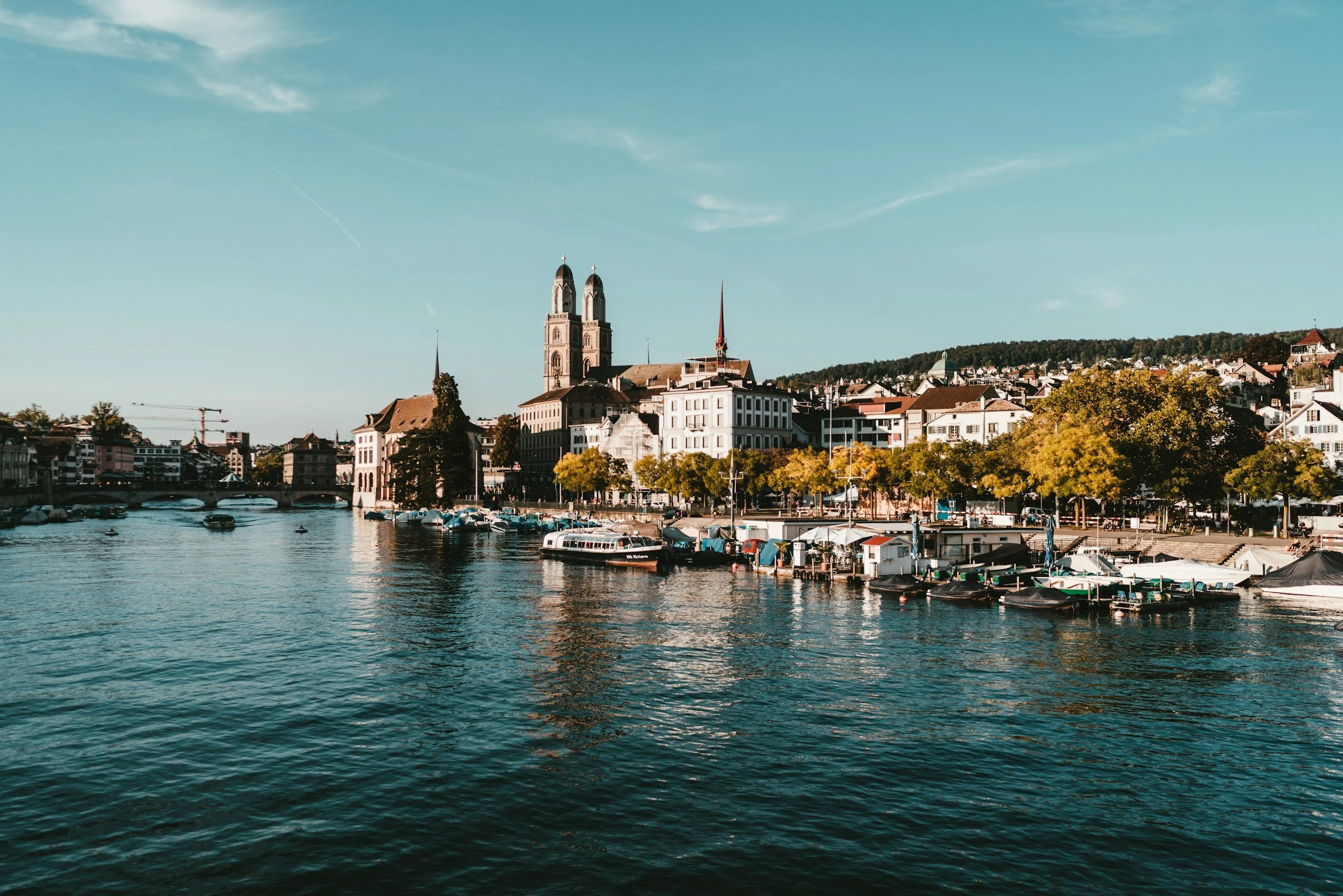 View of a city skyline with a river in the foreground, boats docked along the waterfront, and historic church steeples and modern buildings against a partly cloudy sky.