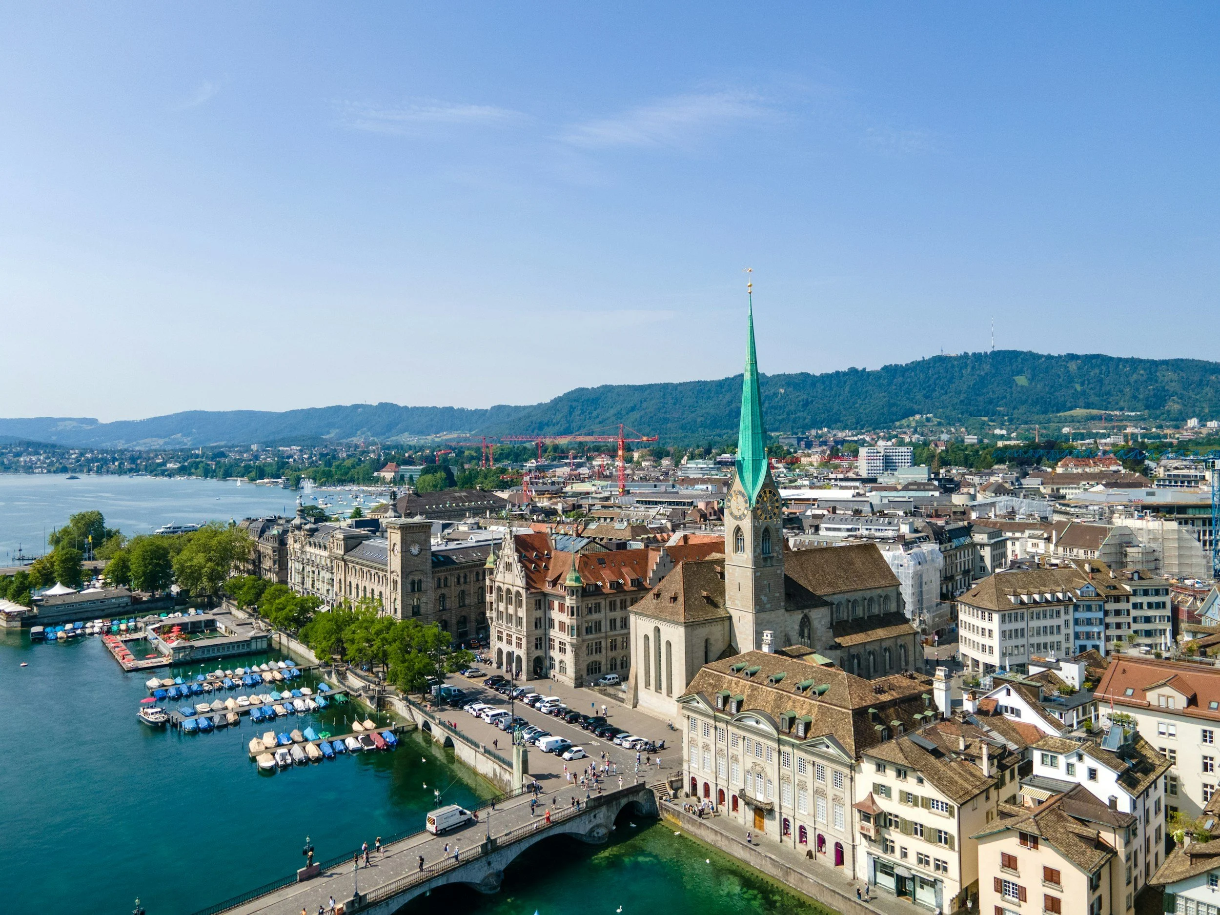 Aerial view of a city with a church featuring a tall green spire, a river with docked boats, and a hillside in the background under a clear blue sky.