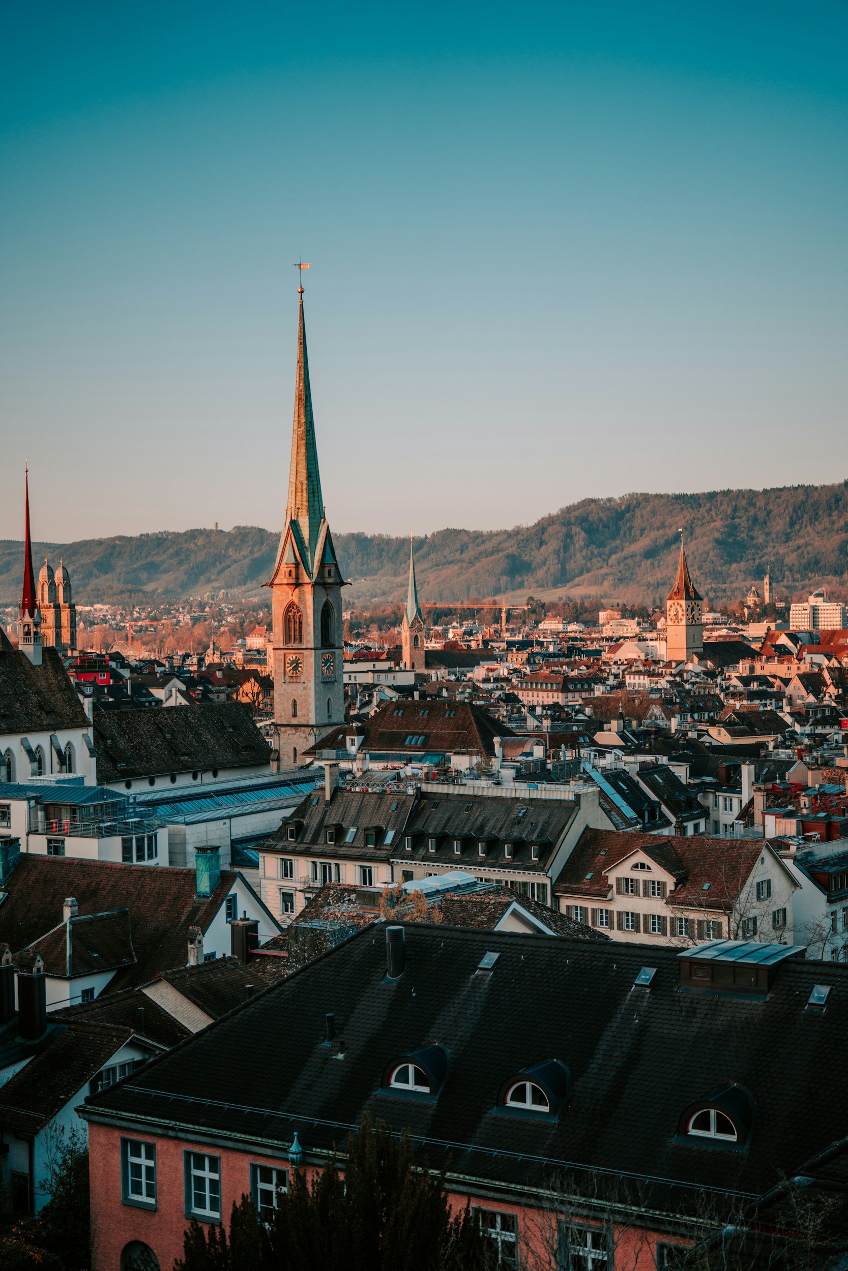 Cityscape of a European town with church steeples, surrounded by hills, during sunset with warm lighting.