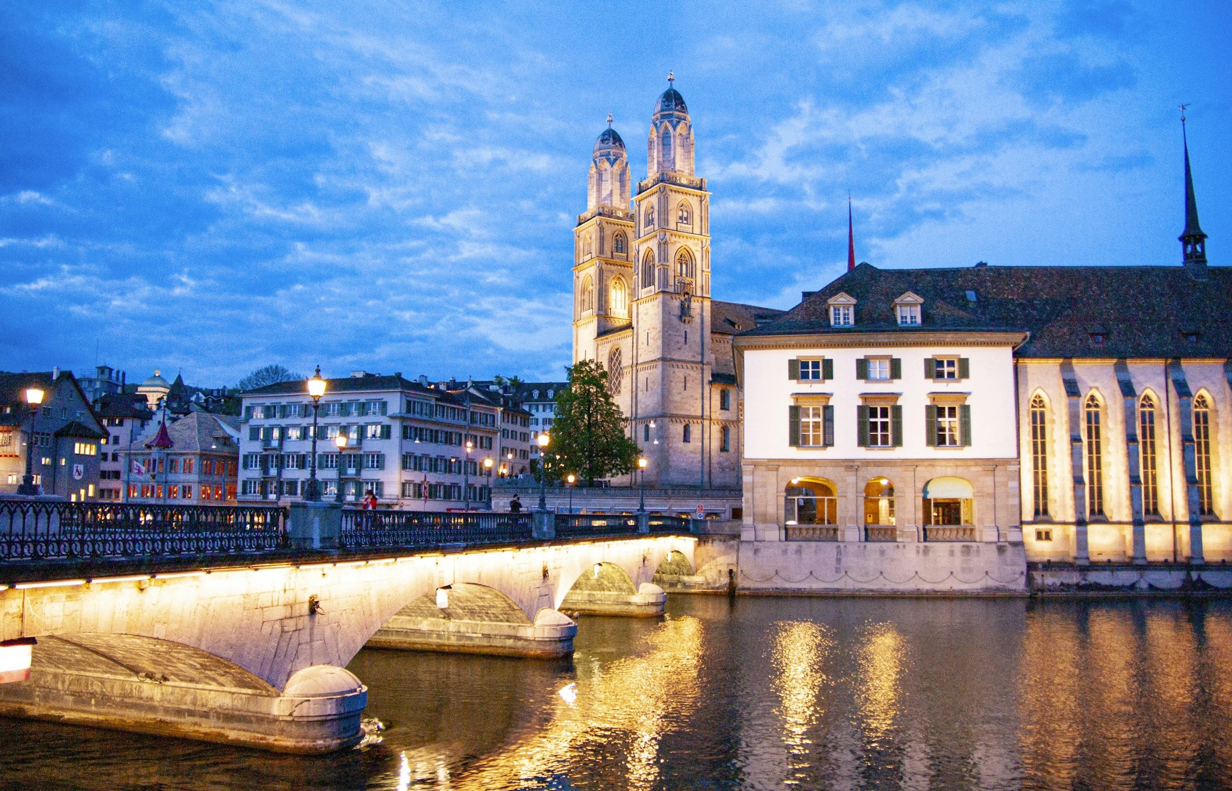 This is a cityscape at dusk with a church and surrounding buildings. There is a bridge over a river reflecting the city lights. The sky is partly cloudy with a blue hue.