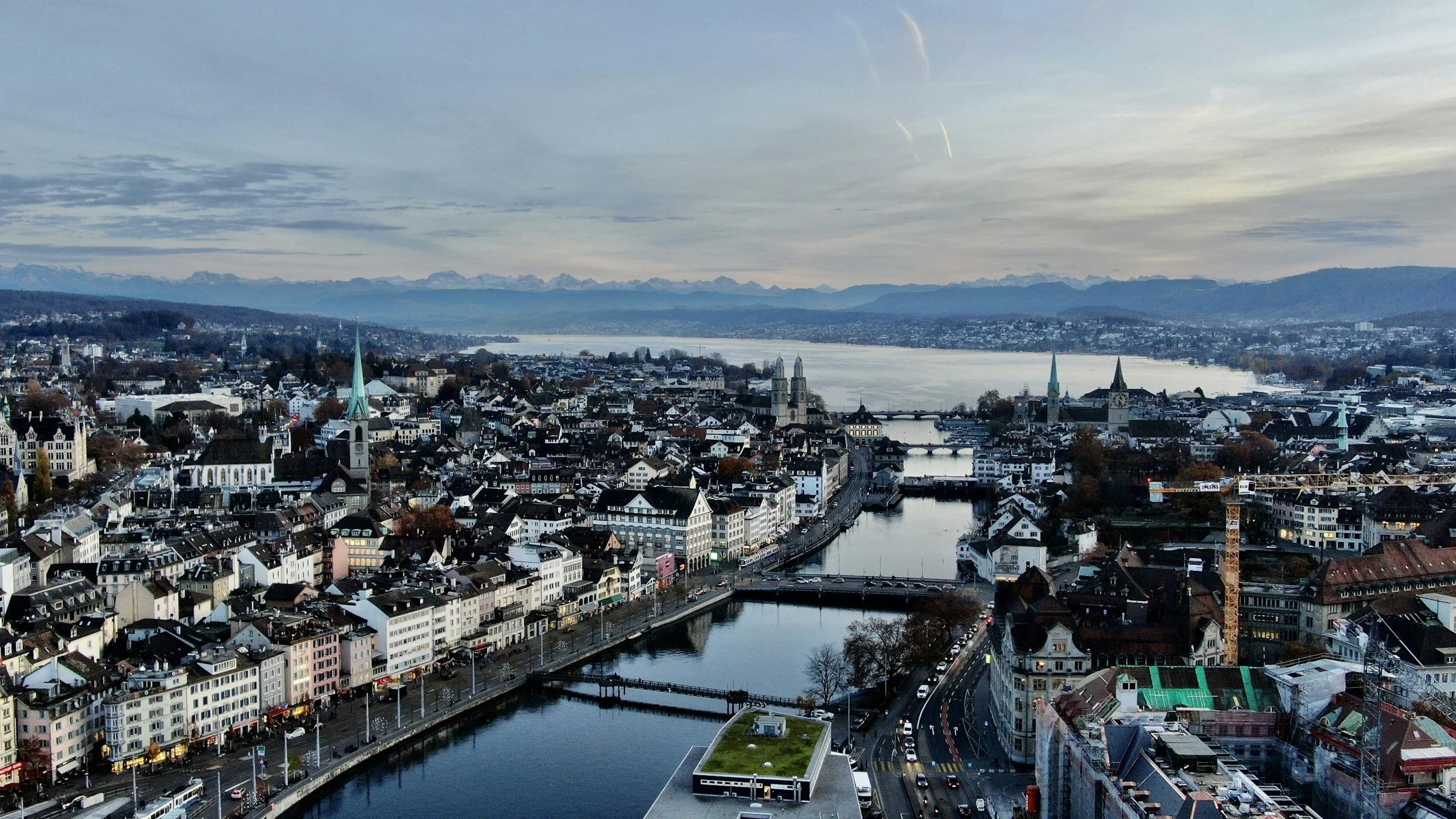 A panoramic view of a city with a river running through it, several church steeples, and mountains in the background under a cloudy sky.
