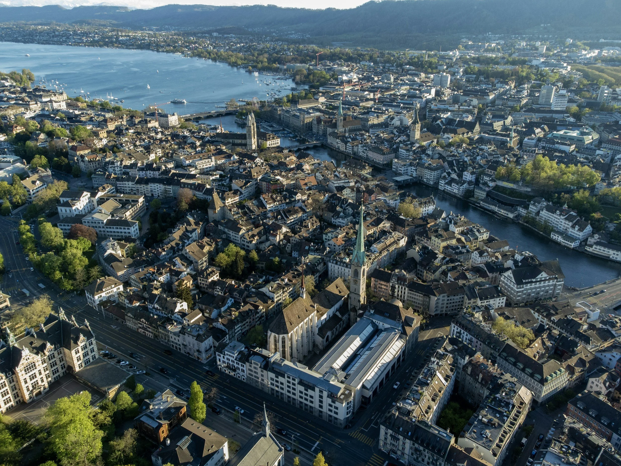 An aerial view of a city near a large body of water, with numerous boats on the water and a mix of historic and modern buildings. Green trees line some streets and parks.