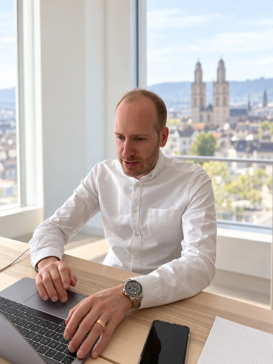 Man in white shirt working on a laptop at a desk, with a smartphone and notebook nearby and a city church visible through the window.