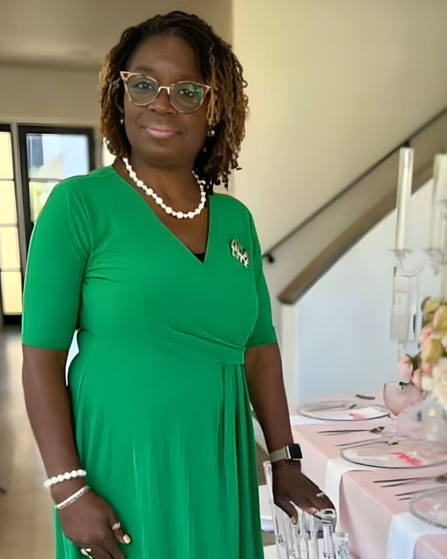 A woman with glasses, wearing a green dress, pearl necklace, and jewelry, standing beside a table with plates and pink flowers, in a well-lit indoor setting.