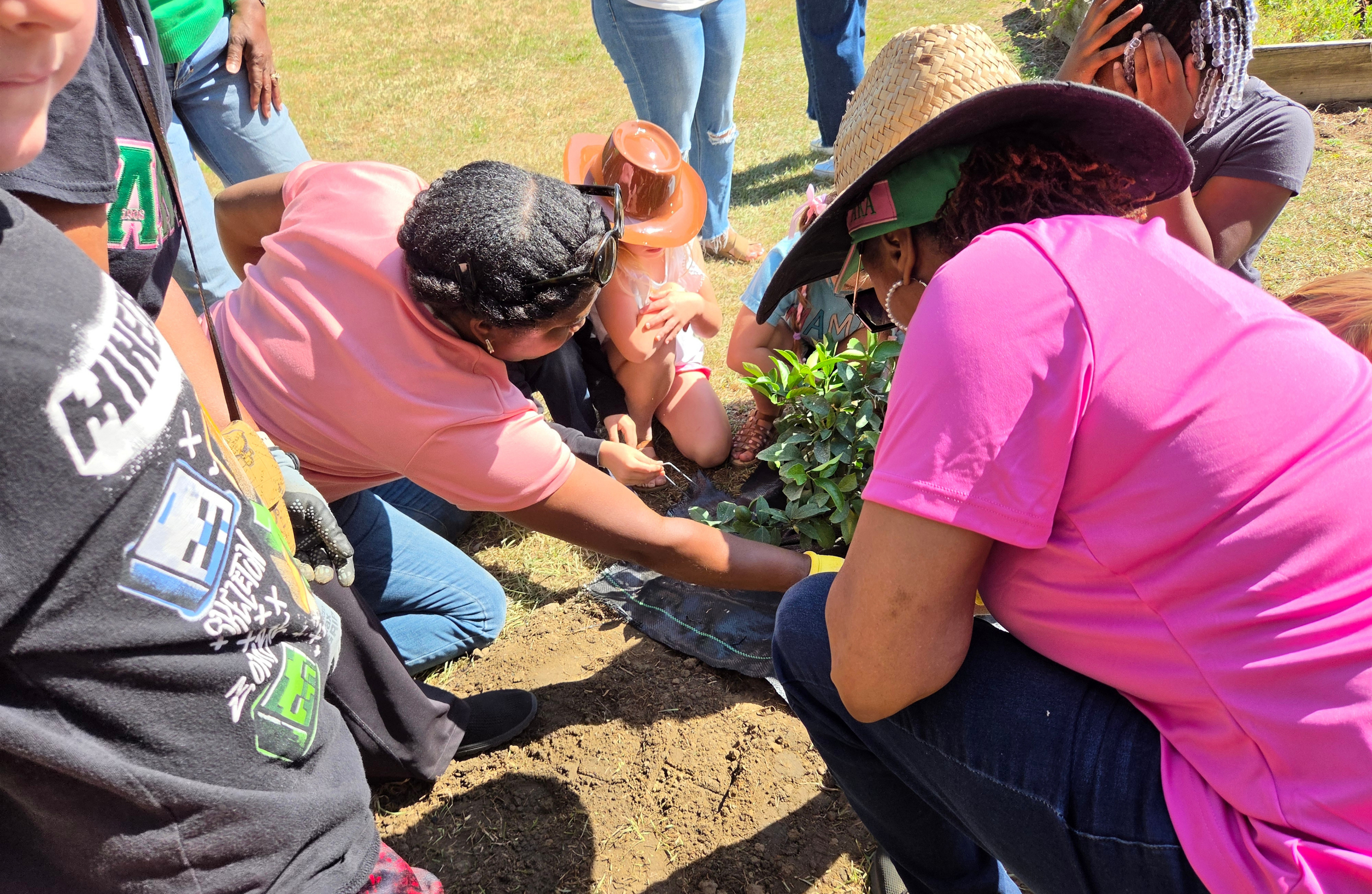 Four women outdoors in a green, natural setting, standing close together, smiling, holding gardening tools, and wearing colorful T-shirts with Greek letters.