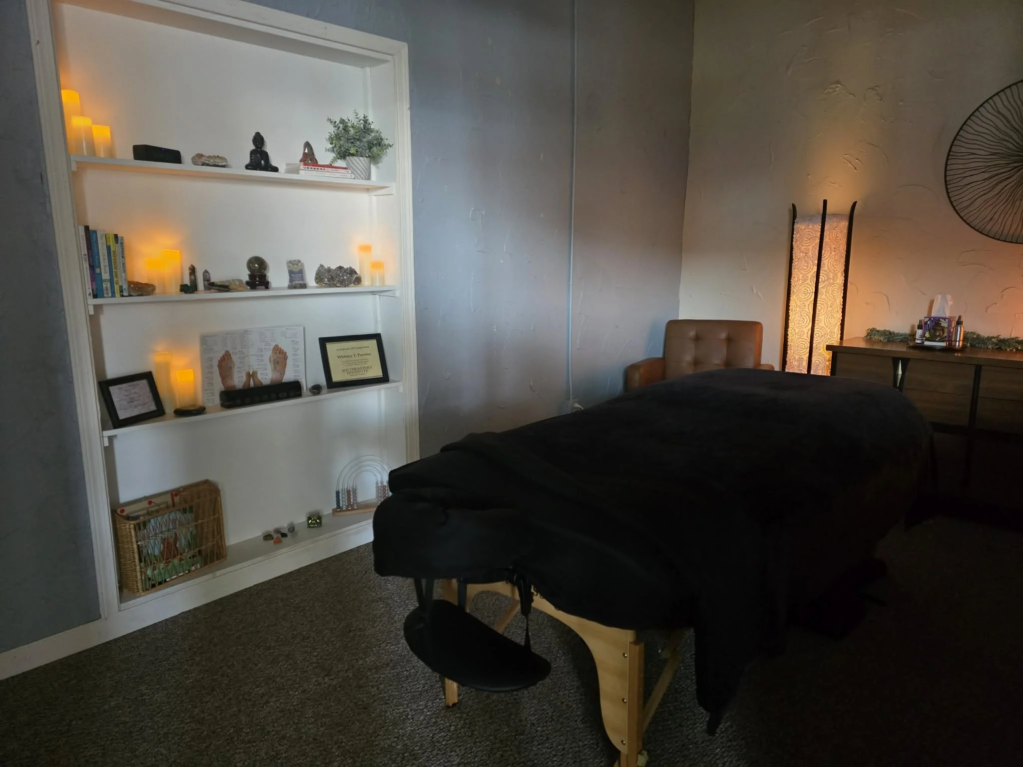 Interior of a massage therapy room with a massage table draped in black cloth, a brown armchair, a white wall with shelves decorated with candles, crystals, and framed certificates, a tall floor lamp, and a wooden dresser with decorative items and tissues.