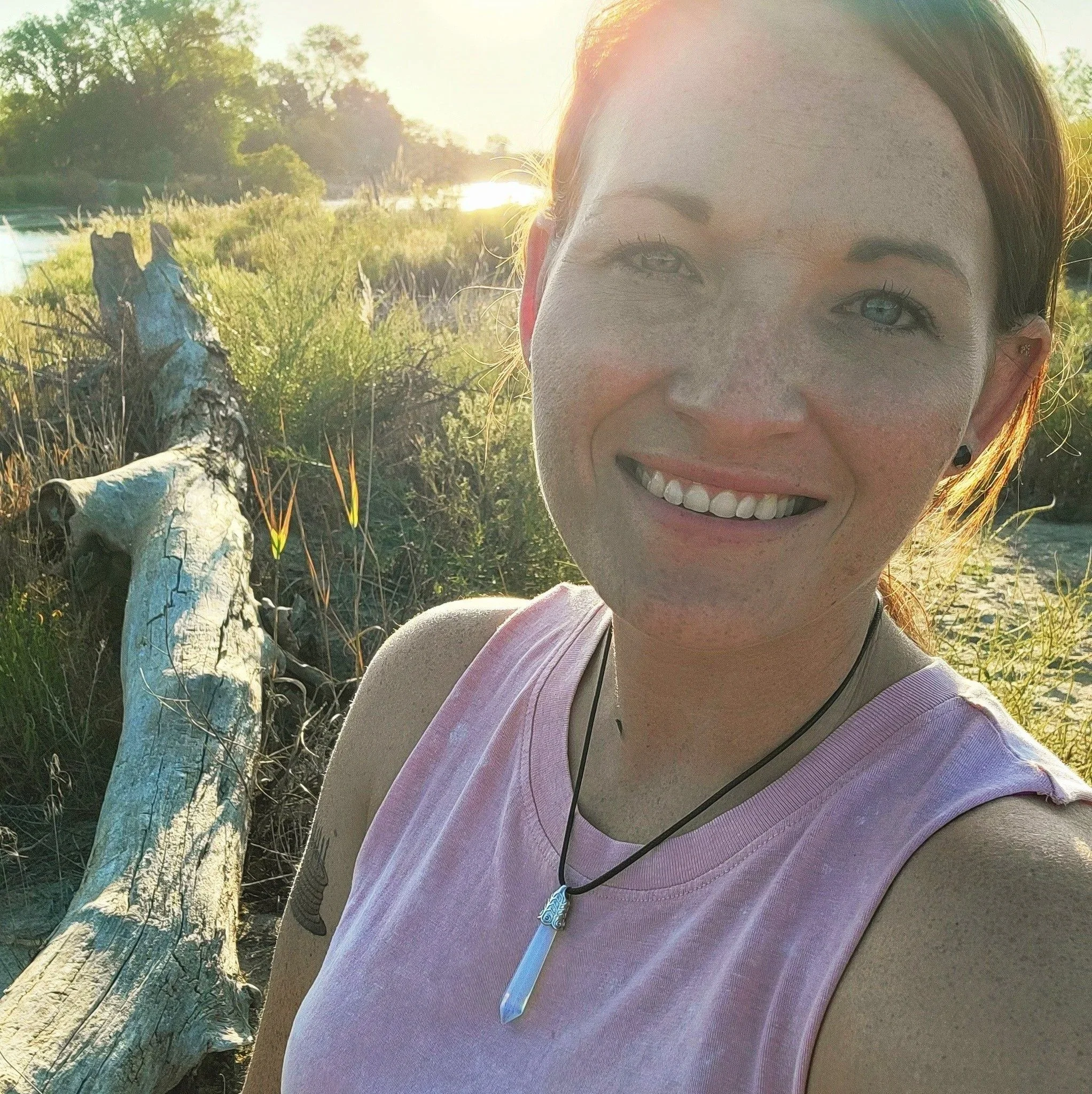 A smiling woman with red hair and freckles taking a selfie outdoors during sunset. She is wearing a pink sleeveless top and a necklace with a pointed pendant. There is a large weathered piece of driftwood and green vegetation in the background.