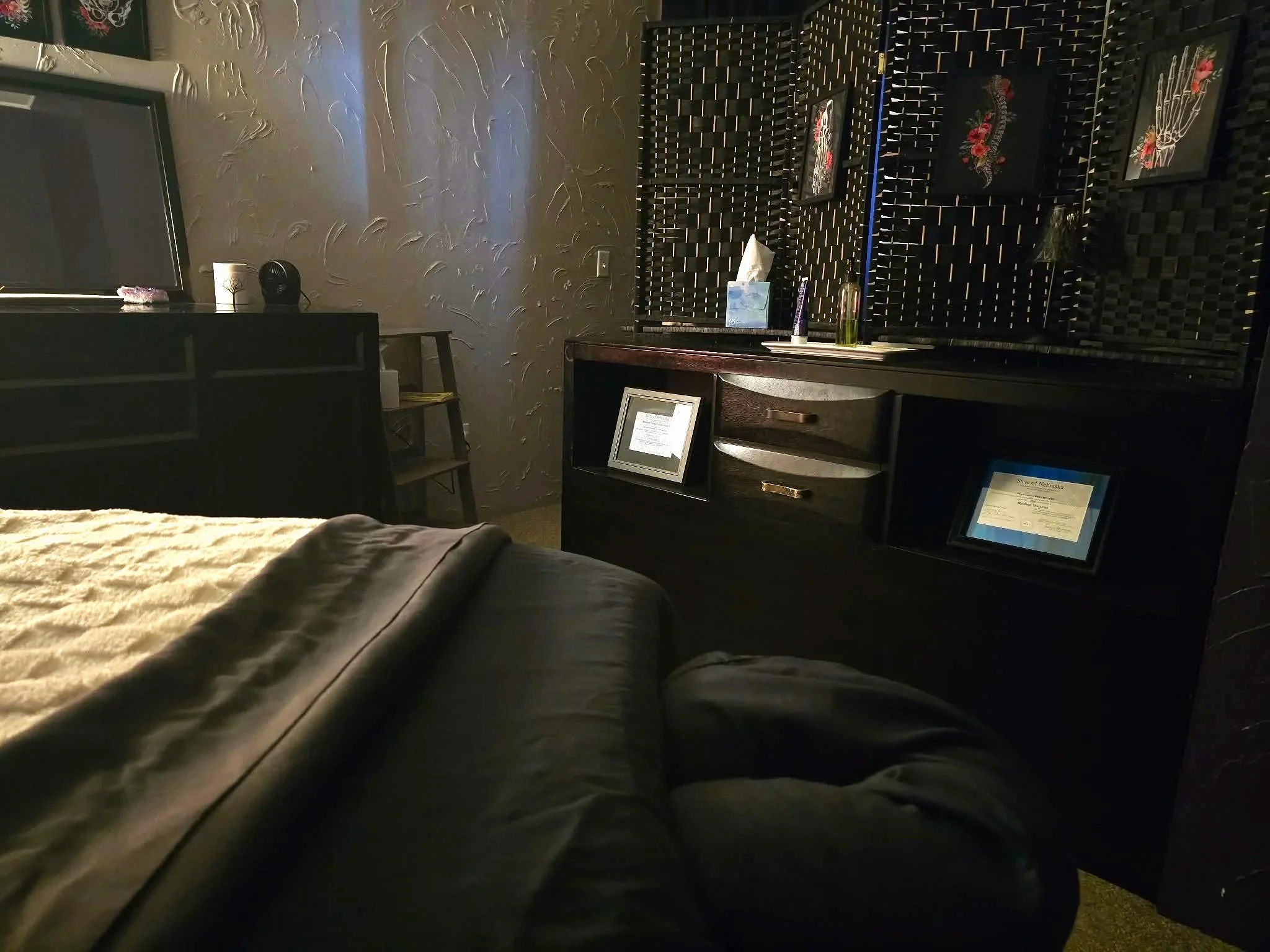 A dark-colored bed with a comforter in the foreground, a black dresser with framed certificates and decorative items on top, and a textured wall with framed artwork in the background.