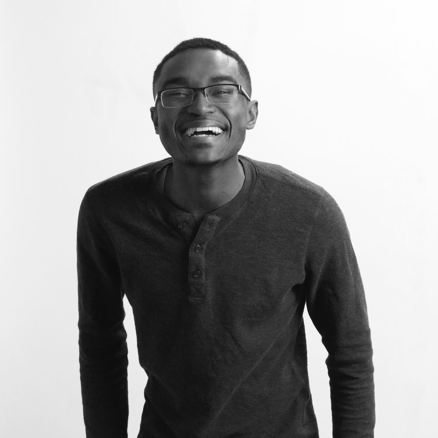 A young man wearing glasses, smiling and laughing, dressed in a casual long-sleeve shirt, standing against a plain white wall.
