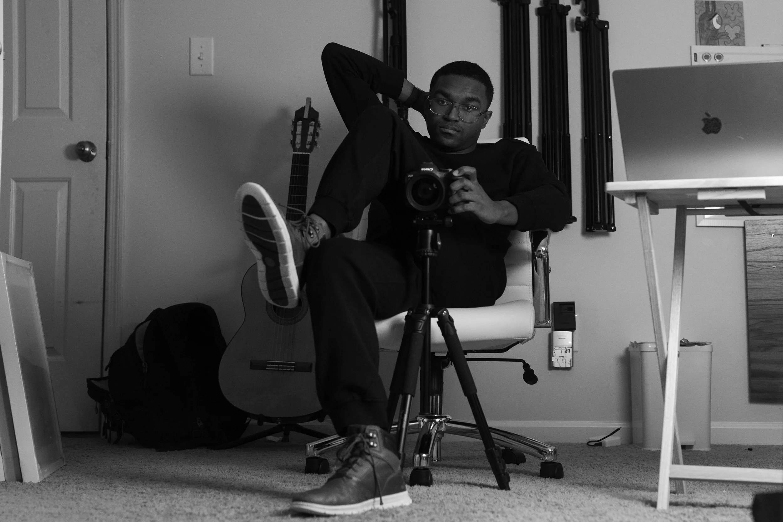 A young man sitting on an office chair, holding a camera, with one leg crossed over the other. He is wearing glasses and sneakers, and is looking directly at the camera. The room has a guitar leaning against the wall, a backpack on the floor, a computer on a desk, and various photographic equipment.