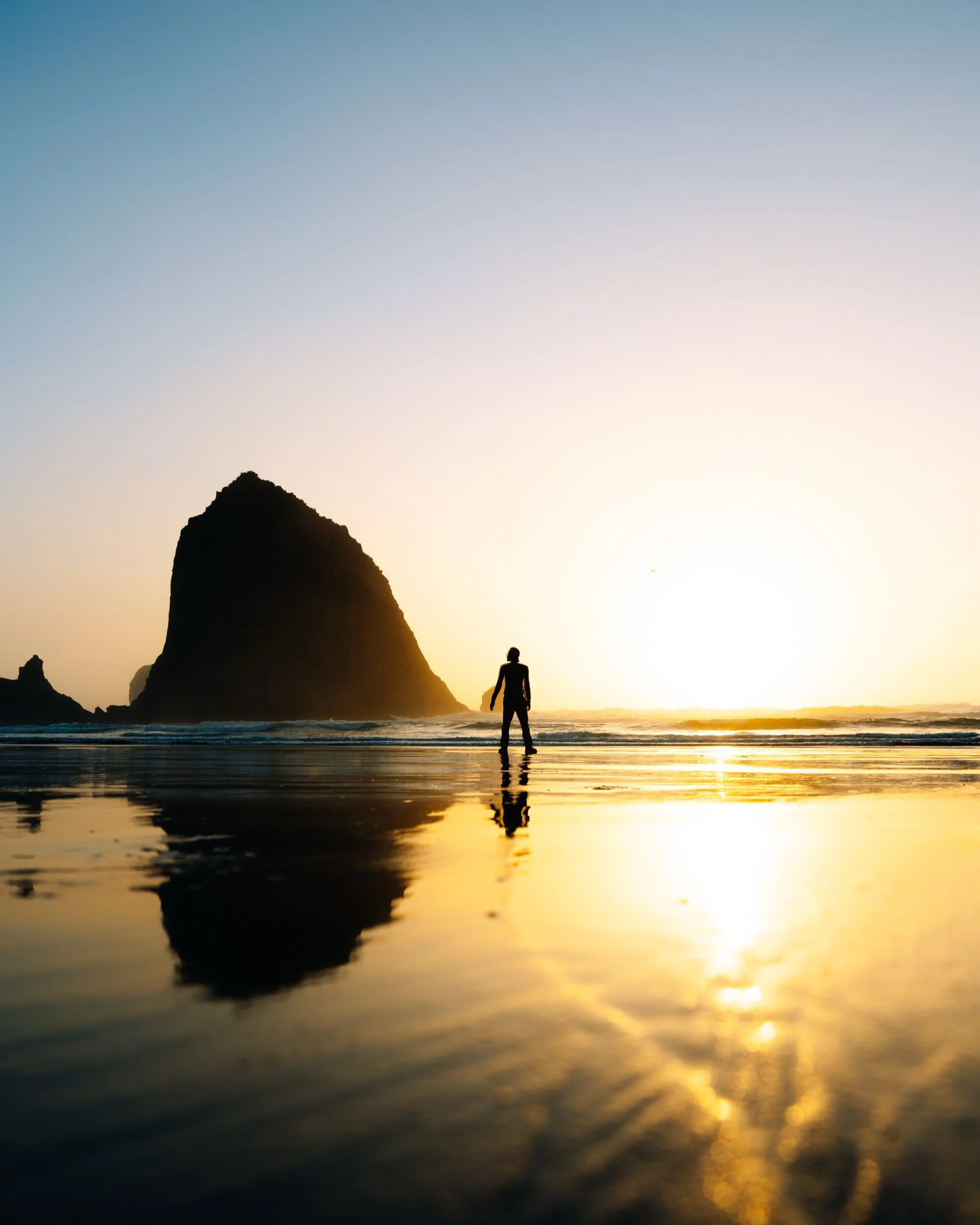 A person standing on the beach at sunset with the ocean and a large rock formation in the background, reflected in the wet sand.