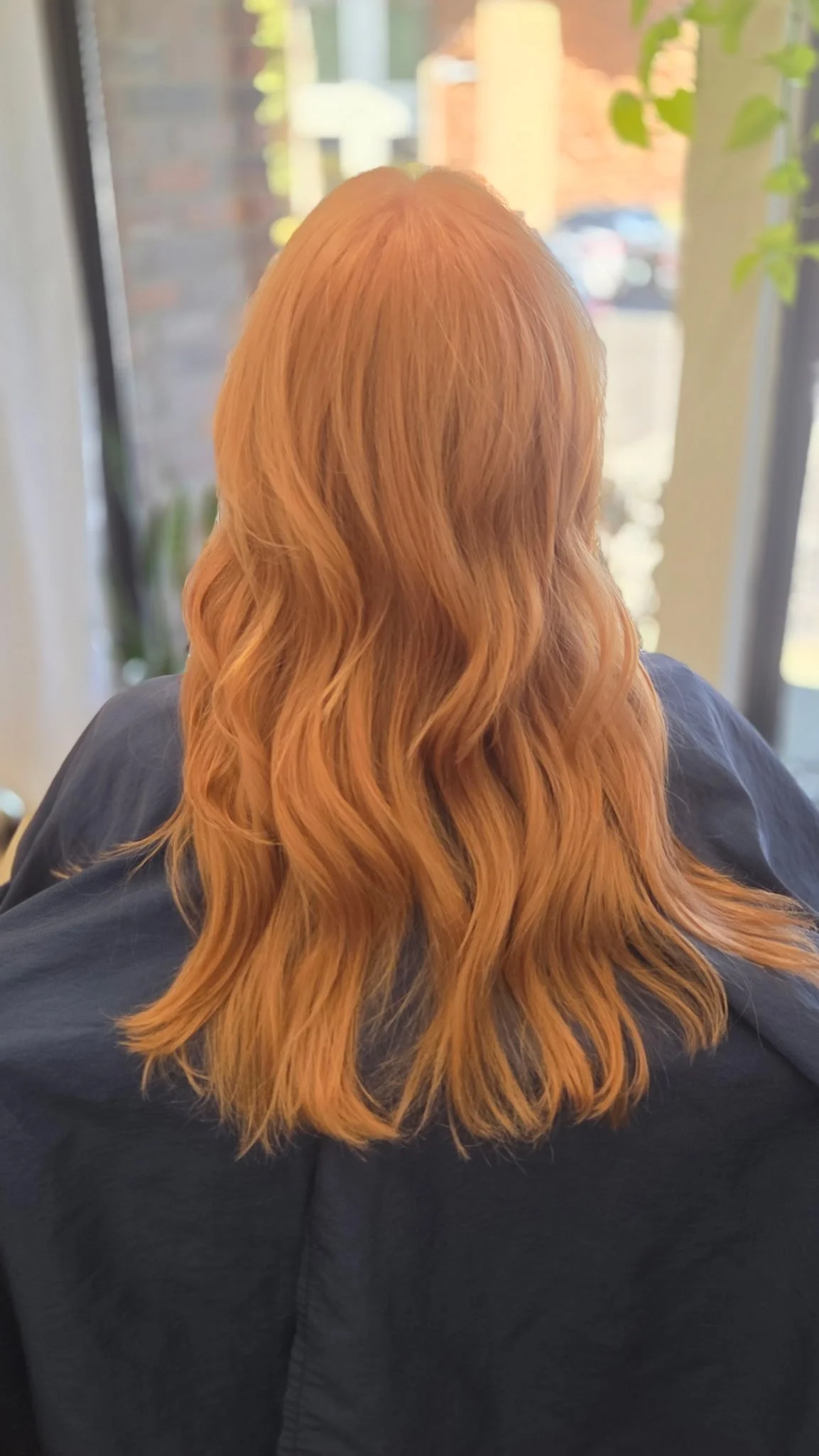Back view of a person with long, wavy red hair sitting indoors near a window.