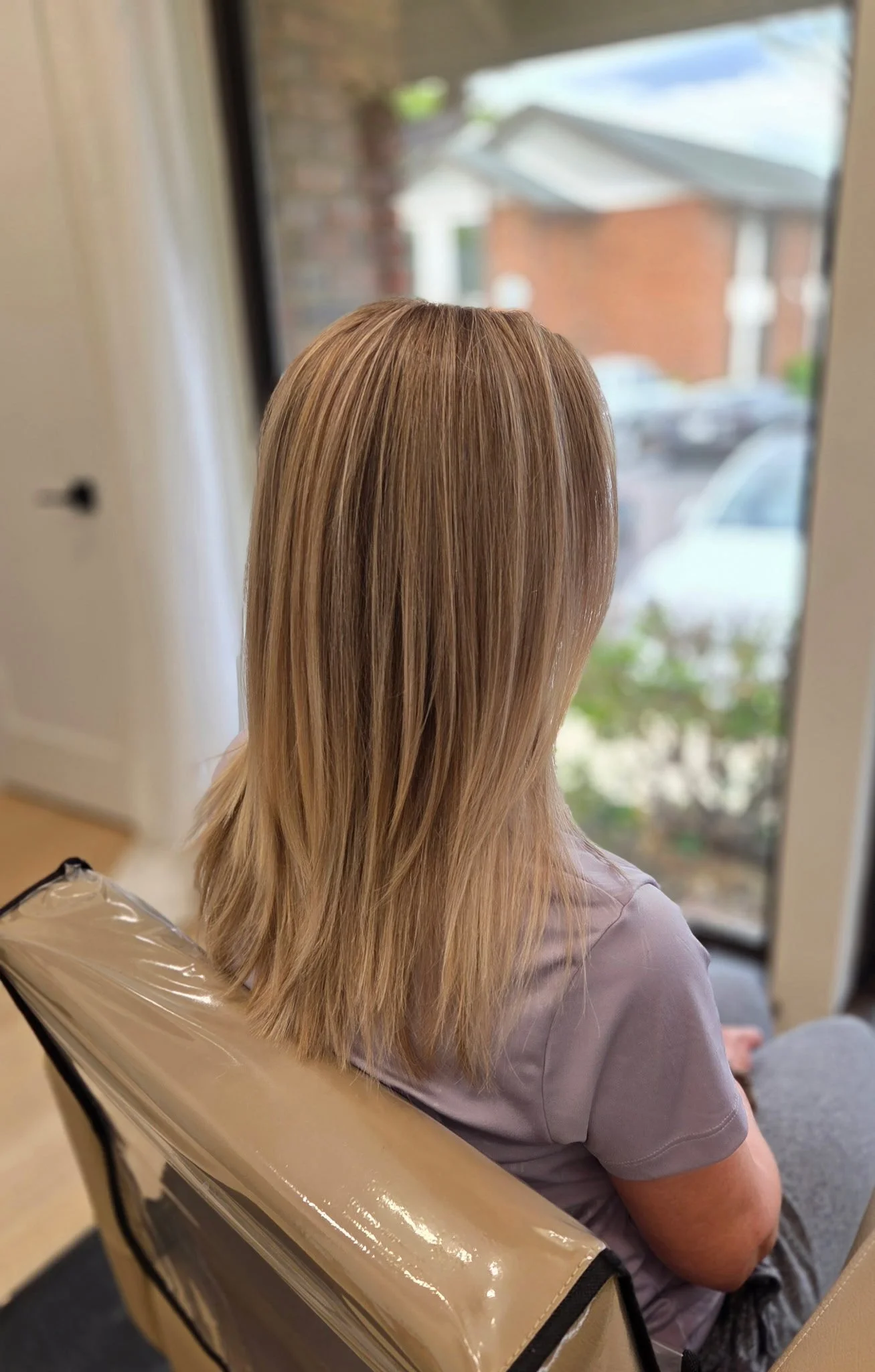 A woman with long blond hair sitting near a window, with a plastic-covered item on her lap, possibly a hair or beauty treatment.