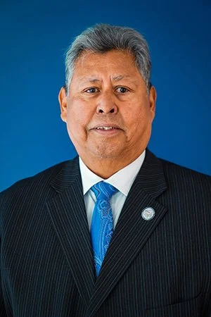 Portrait of a middle-aged man in a black suit with a blue tie, standing against a blue background.