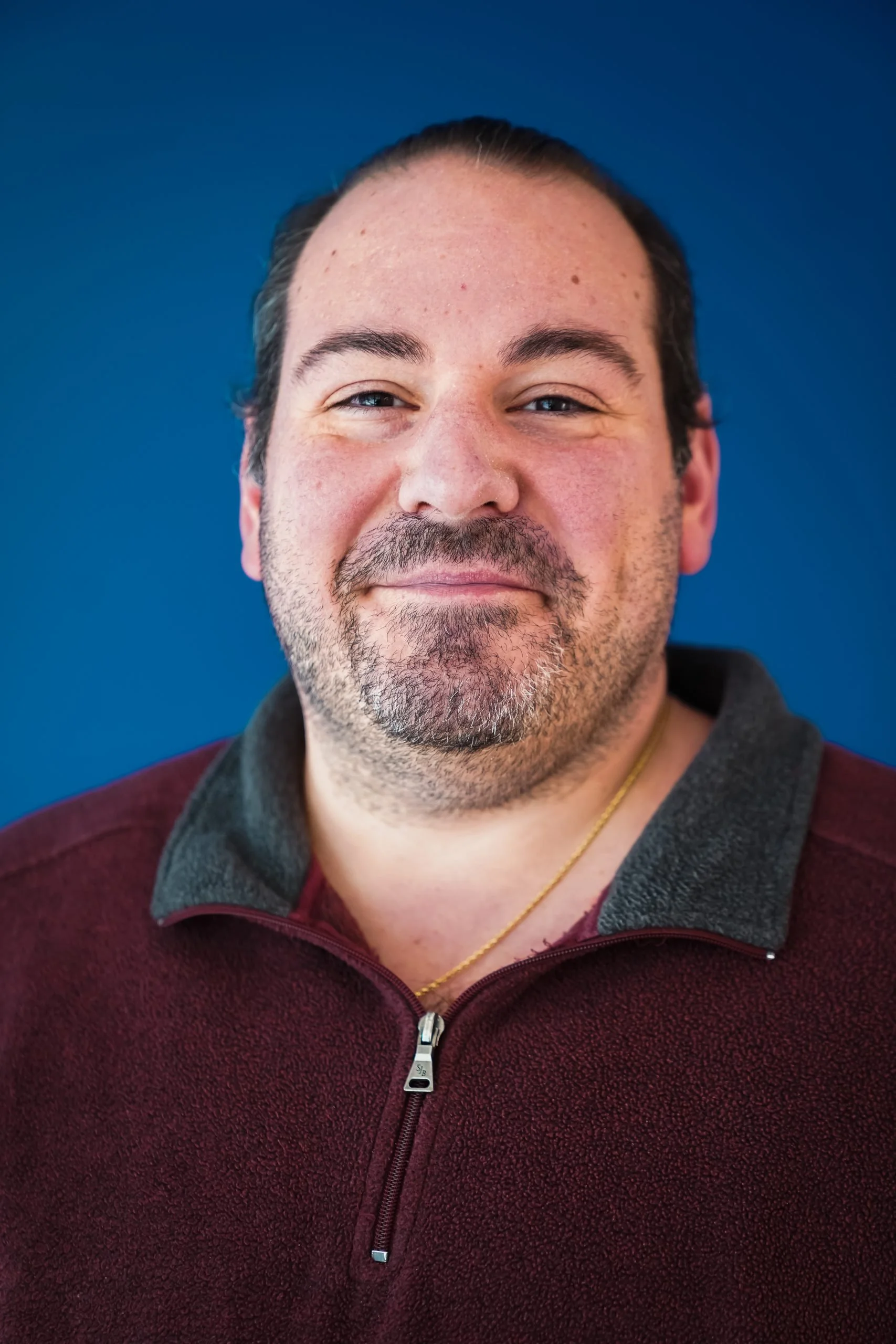 Close-up of a smiling man with dark hair, stubble, and a mustache, wearing a maroon fleece jacket with a zip-up front and a gold chain, standing against a solid blue background.