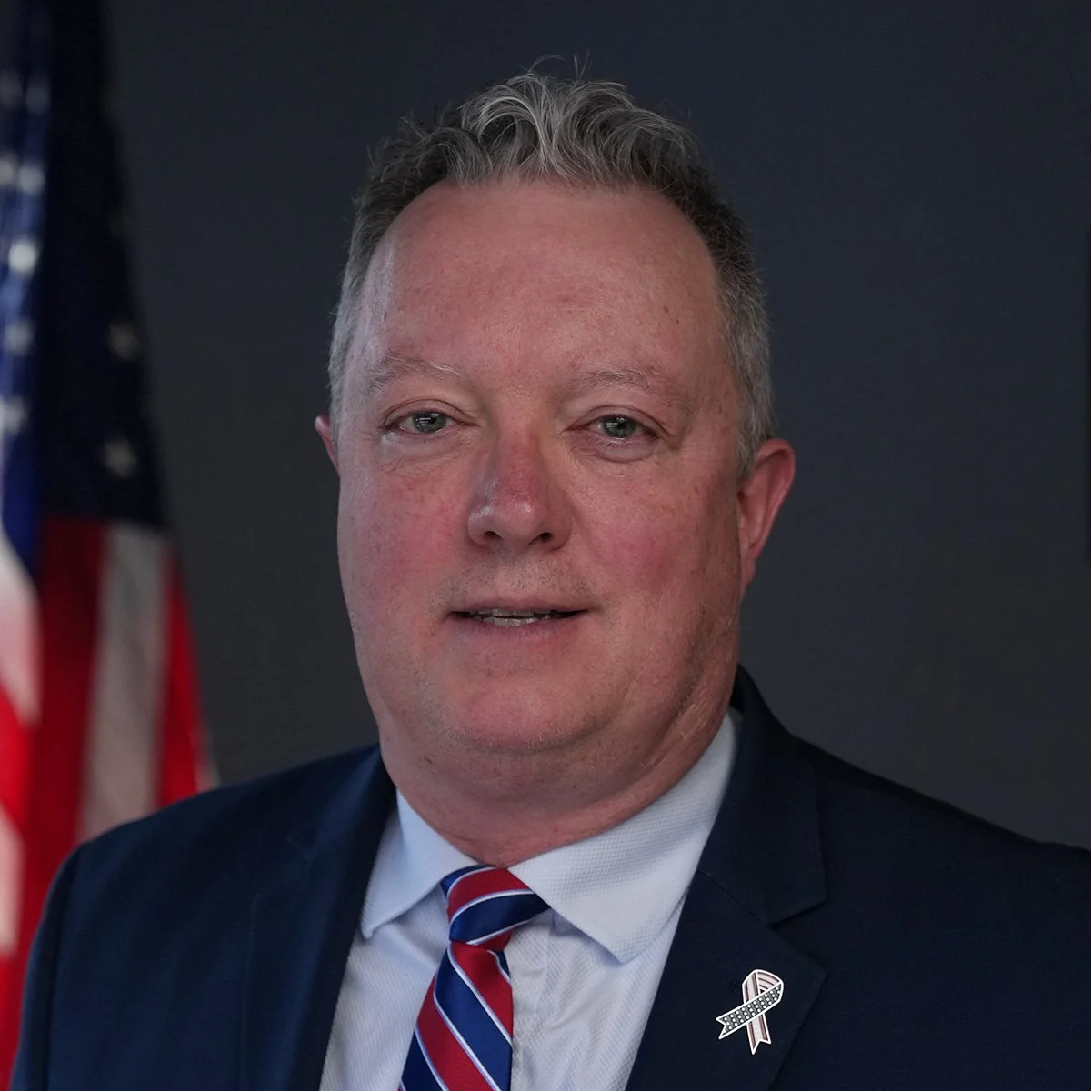 A man with light skin, short blonde hair, wearing a dark suit, white shirt, and a red, white, and blue striped tie. He has a ribbon lapel pin on his suit. An American flag is partially visible to his left, with a dark background behind him.