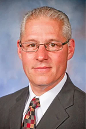 A man with short, gray hair, glasses, and a suit with a patterned tie, posing for a professional headshot against a blue background.
