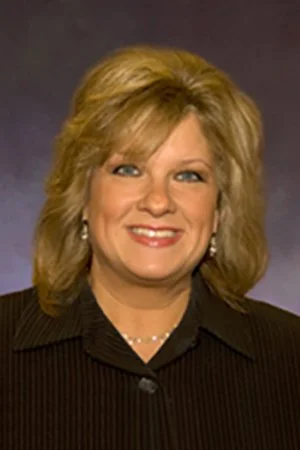 A smiling woman with blonde hair wearing a black blazer and pearl earrings, with a dark gray background.