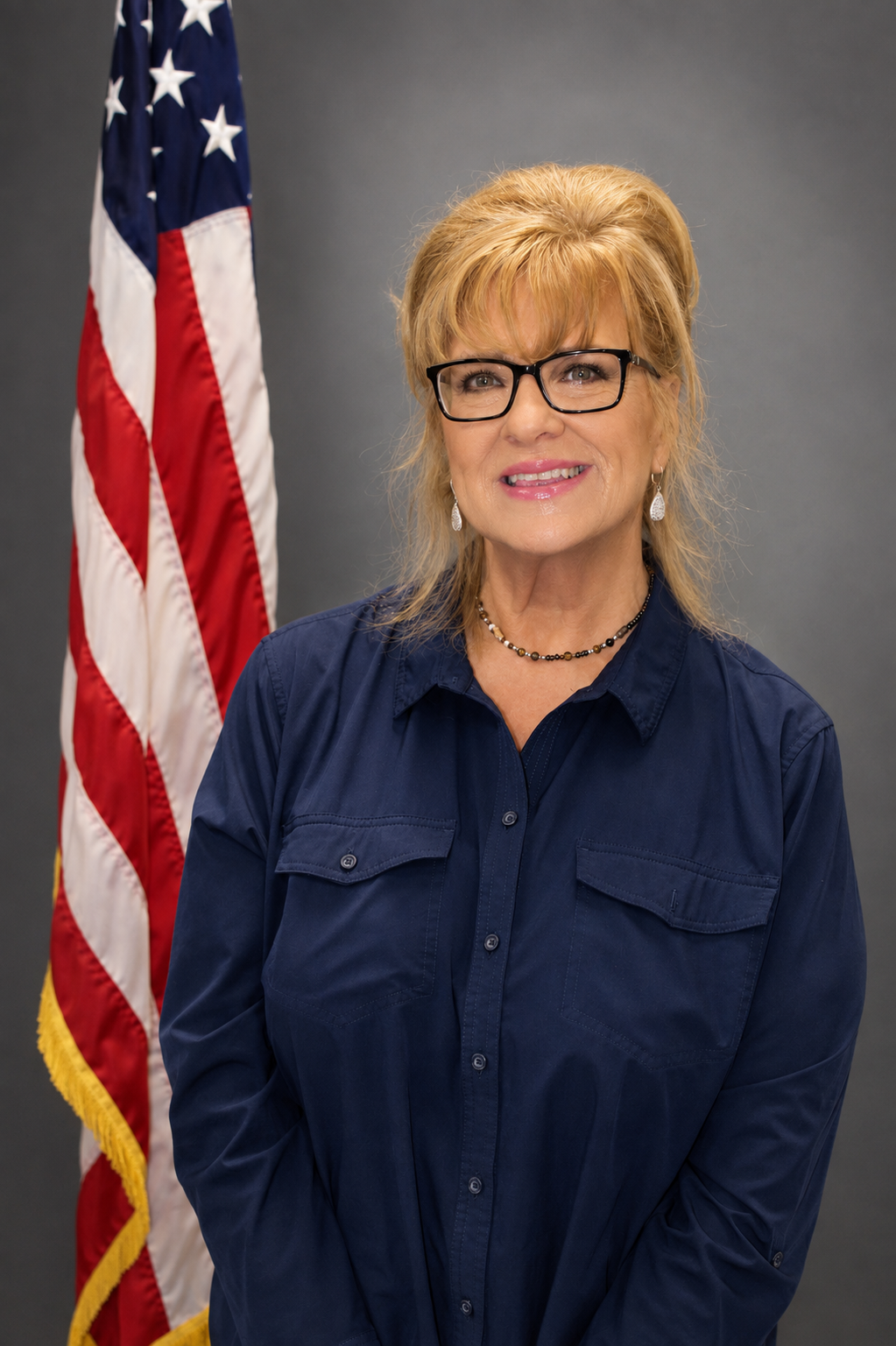 A woman with blonde hair, wearing glasses, a navy blue shirt, and jewelry, smiling at the camera, standing in front of an American flag against a gray background.