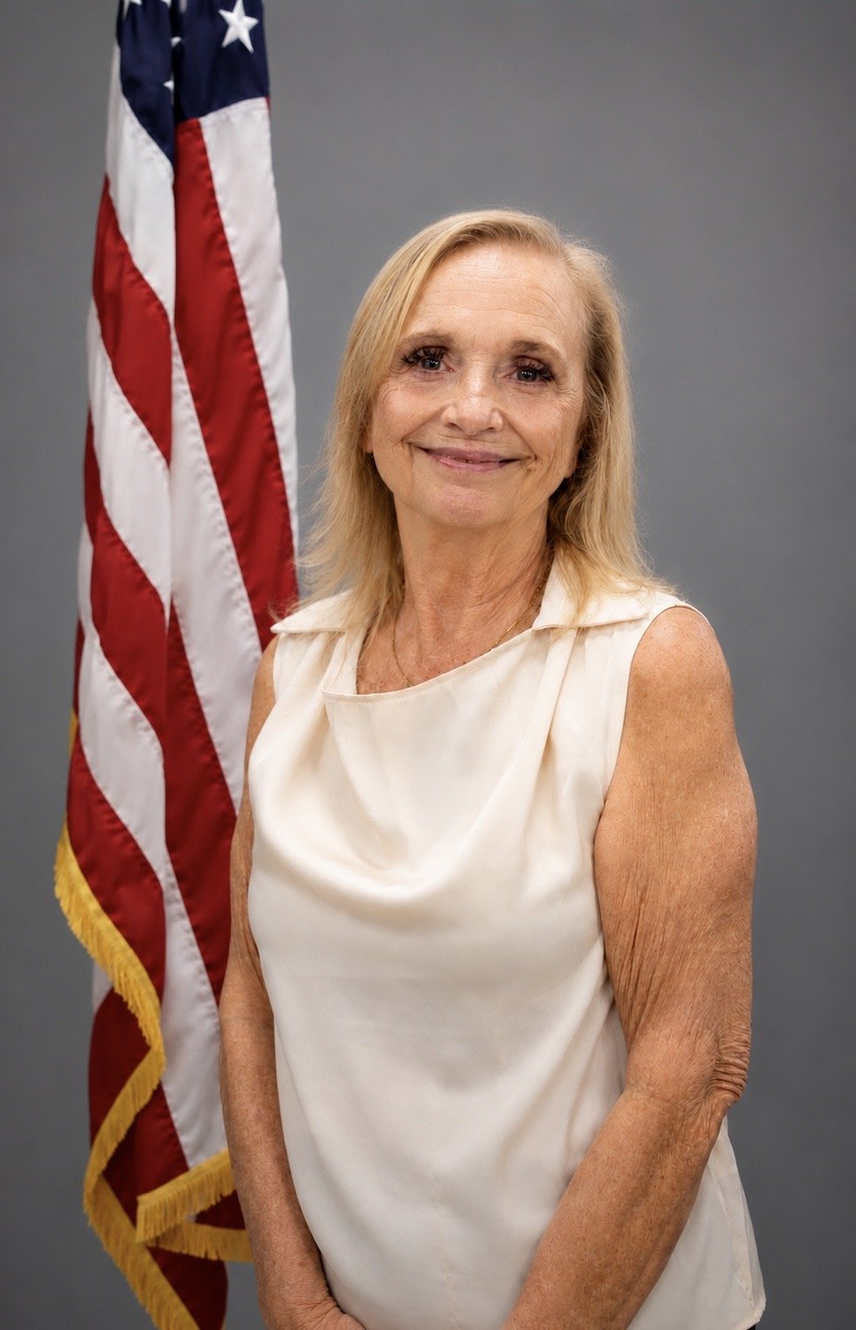 A smiling older woman with blonde hair standing in front of an American flag wearing a sleeveless white top.