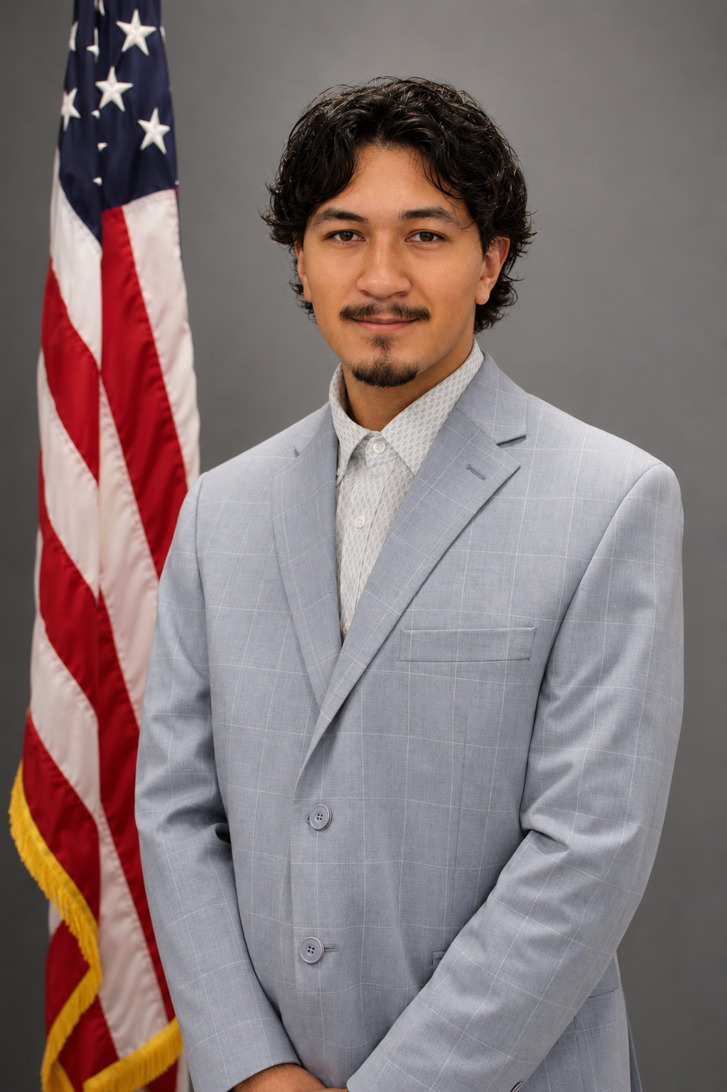 A young man with dark curly hair and a mustache, wearing a light gray checked suit and white patterned shirt, stands in front of an American flag against a gray background.