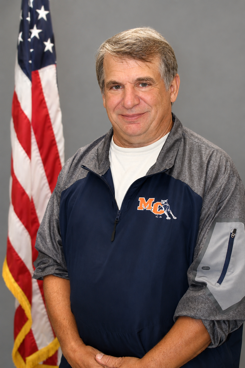 A man standing in front of an American flag, wearing a gray and navy zip-up jacket with a logo that says 'MC' and depicts a dog.