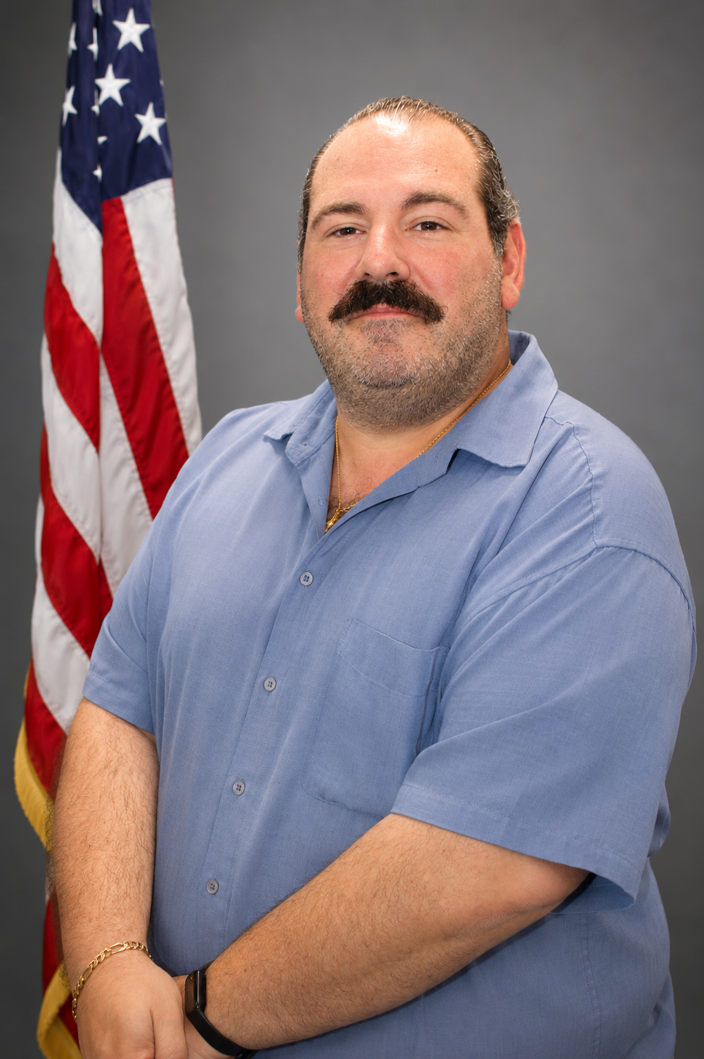 A man with a mustache and beard standing in front of an American flag, wearing a blue short-sleeved shirt.