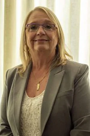 A middle-aged woman with blonde hair and glasses, wearing a gray blazer and white top, standing indoors with light-colored curtains behind her.
