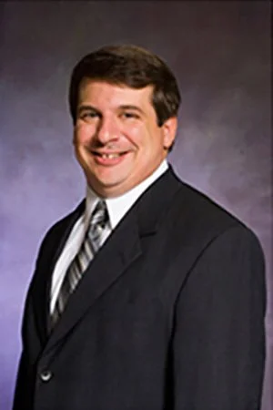 A man in a dark suit with a white shirt and patterned tie, smiling against a dark background.