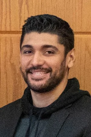 A man with short dark hair and a beard smiling at the camera, wearing a dark hoodie, standing against a wood-paneled wall.