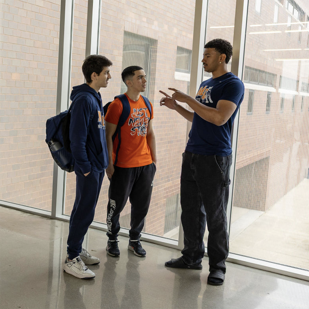 Three young men talking near a glass wall, two are students with backpacks, one is talking with hand gestures, in a school hallway.