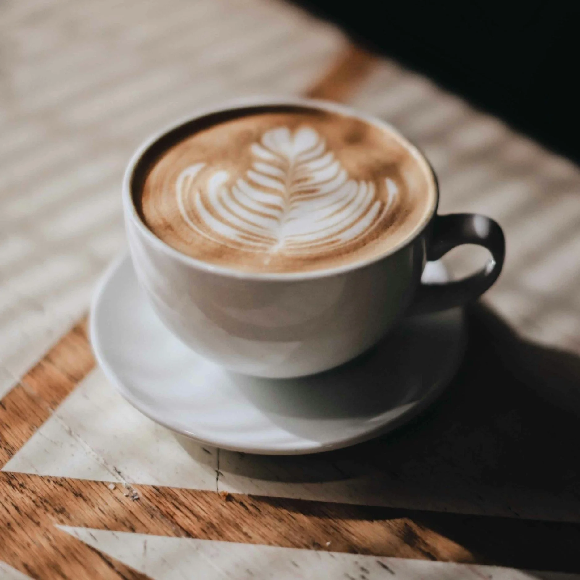 A cup of coffee with latte art resembling a leaf pattern, placed on a saucer on a wooden table.
