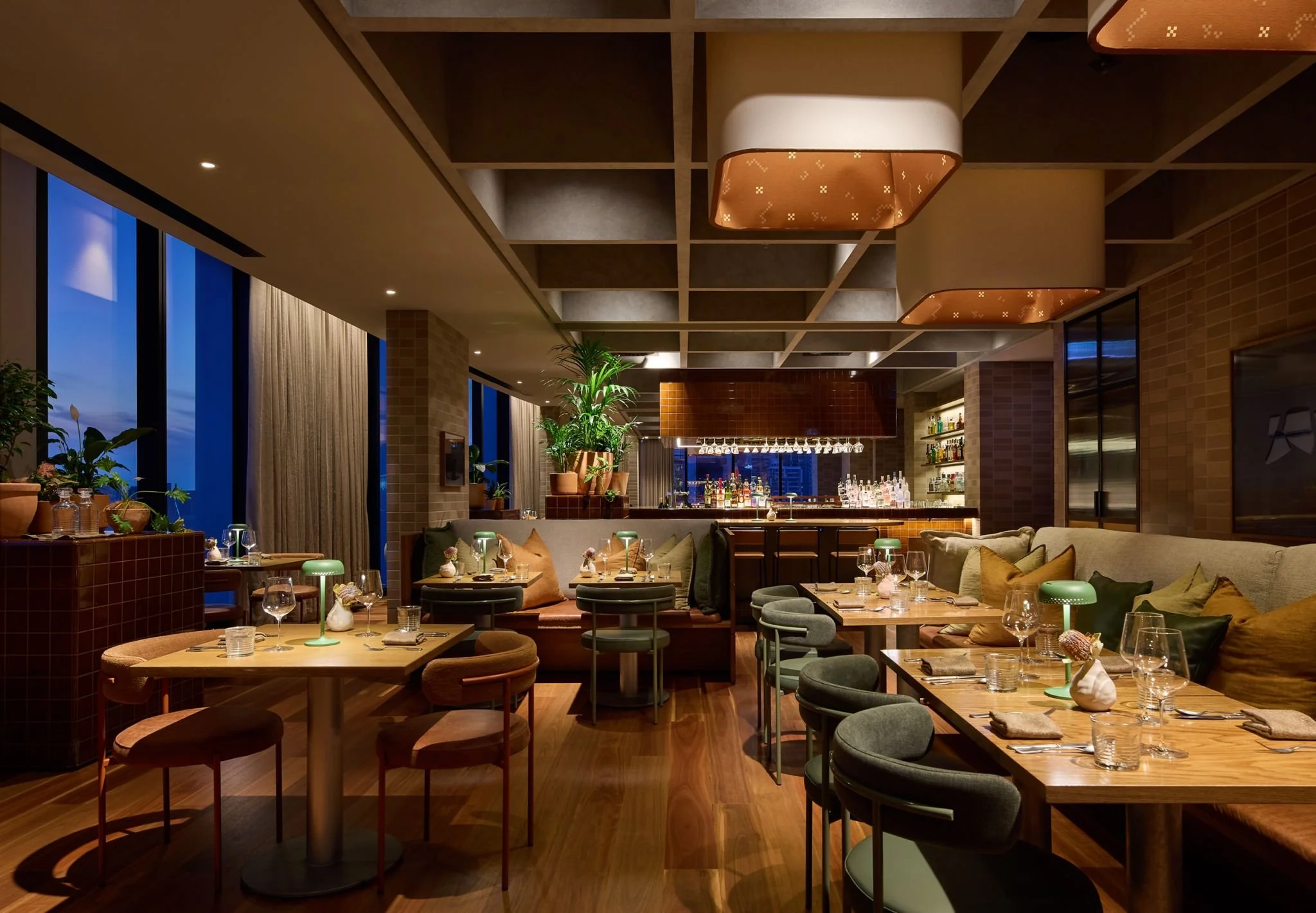 Interior of a modern restaurant with wooden furniture, potted plants, and large windows showing a cityscape at dusk. The ceiling has decorative lighting fixtures, and there is a bar area in the background with hanging glasses.