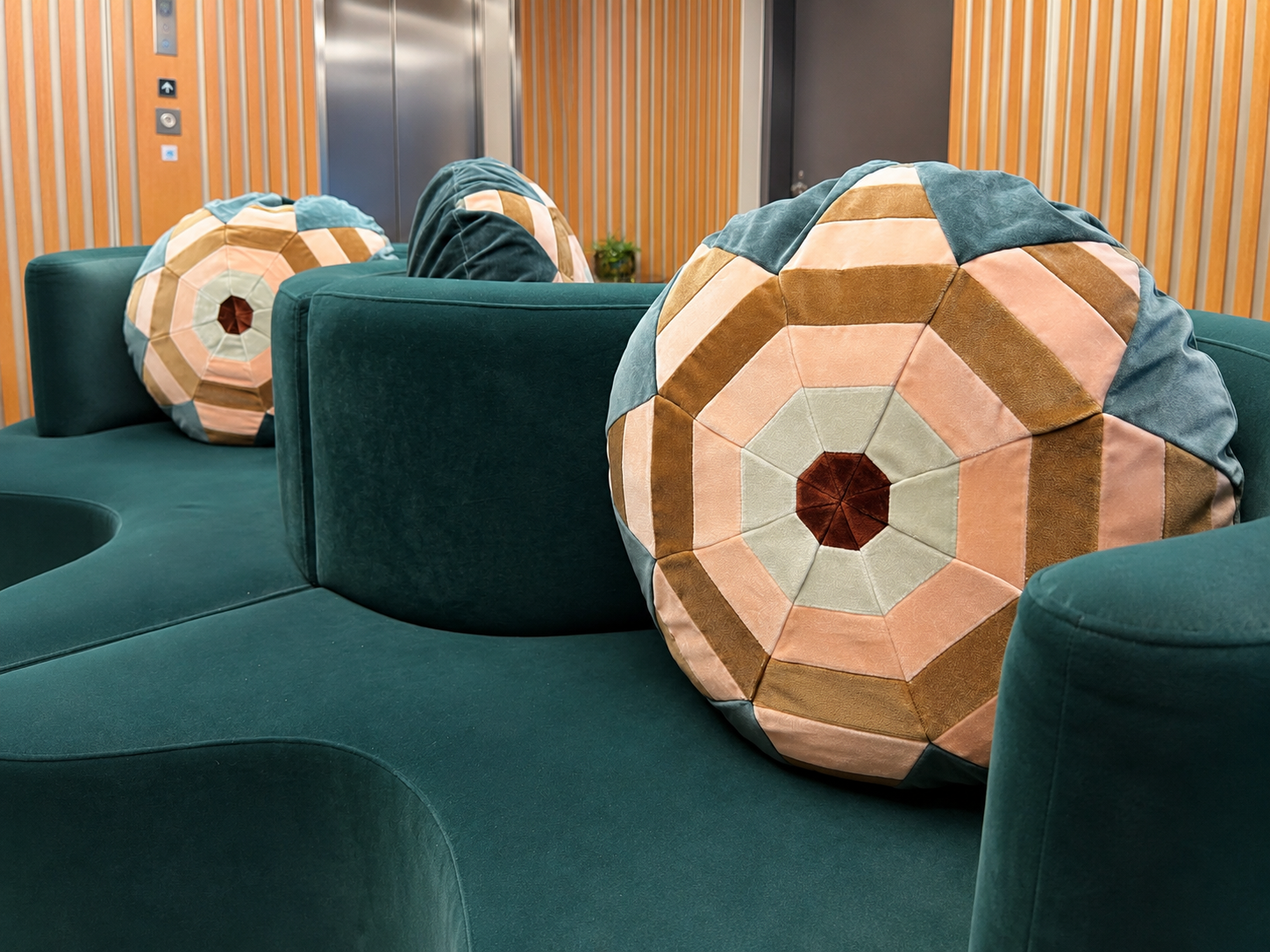 Green sofa with colorful geometric patterned round pillows in an indoor setting with wood panel walls and an elevator in the background.