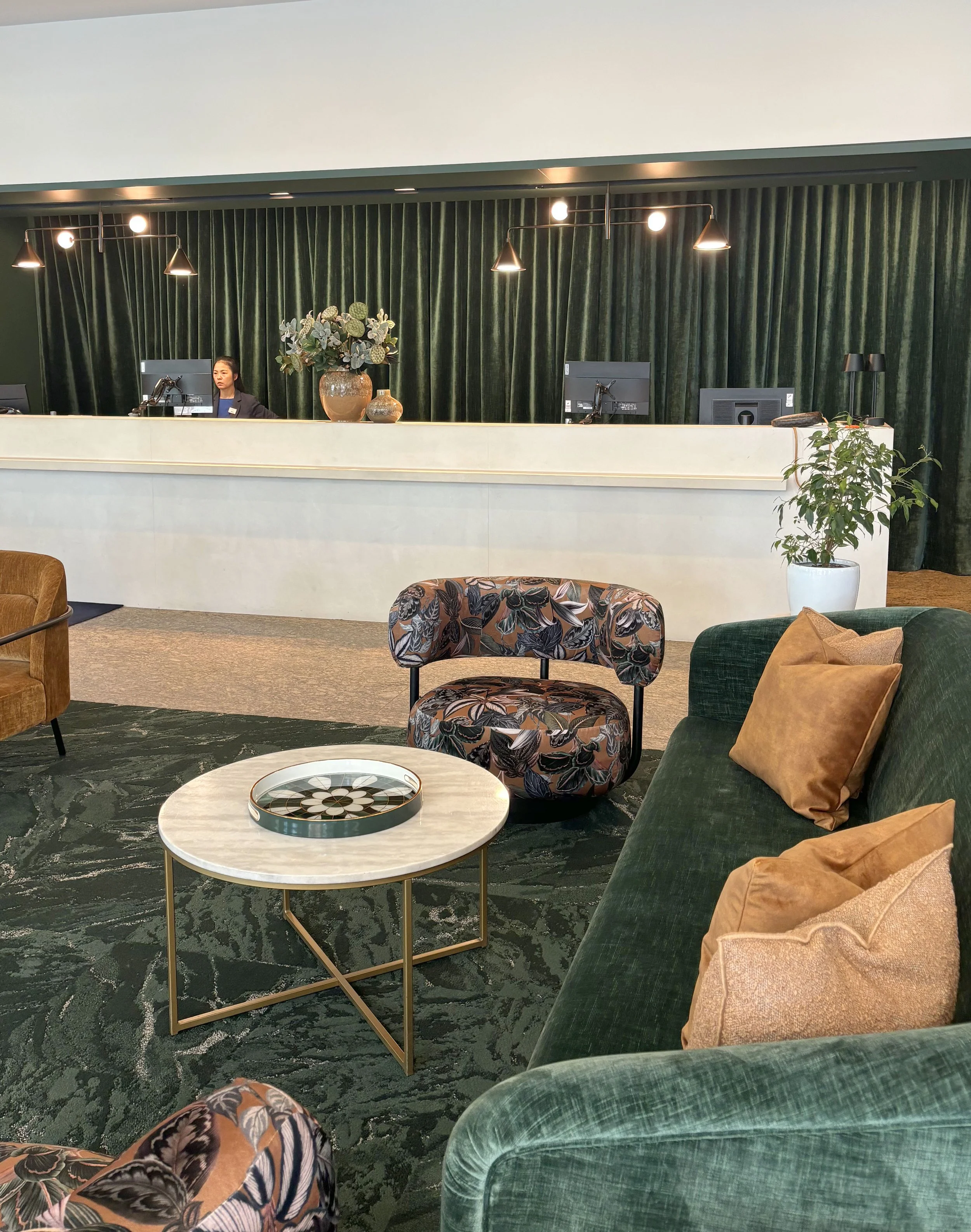 Hotel lobby with green velvet curtains, a reception desk with a woman, floral arrangements, a green sofa with pillows, a round coffee table with a decorative tray, and patterned chairs.