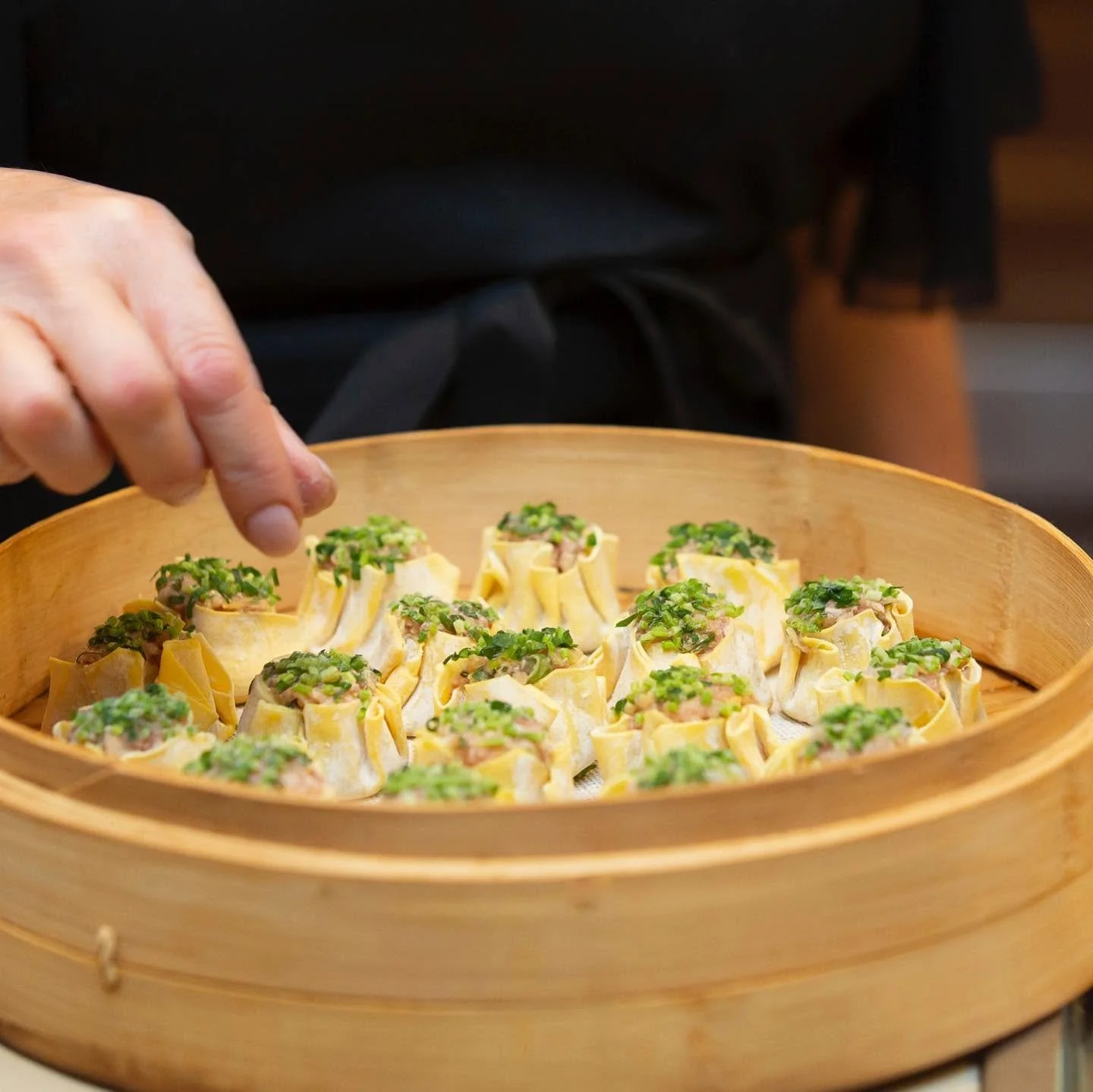 Close-up of a person's hand topped with chopped green onions, placing or adjusting dim sum dumplings in a round bamboo steamer. The dumplings have thin yellow wrappers and are garnished with green onions.