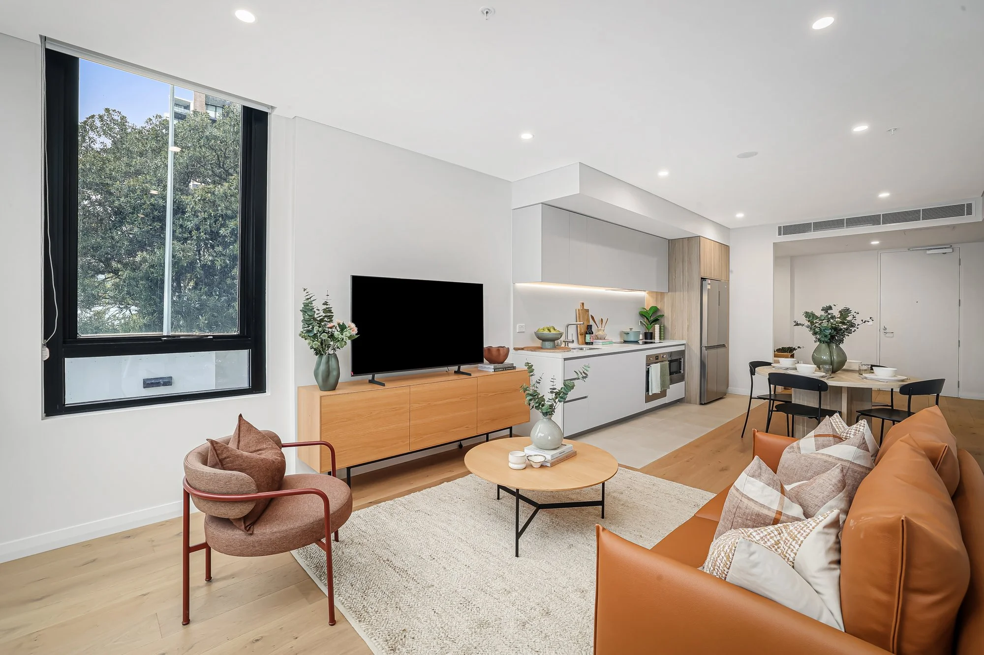 Modern living room with a large window, a brown armchair, a leather sofa with pillows, a round coffee table with a vase, a TV on a wooden console, open concept kitchen with white cabinets, a dining table with a vase and chairs, and indoor plants.