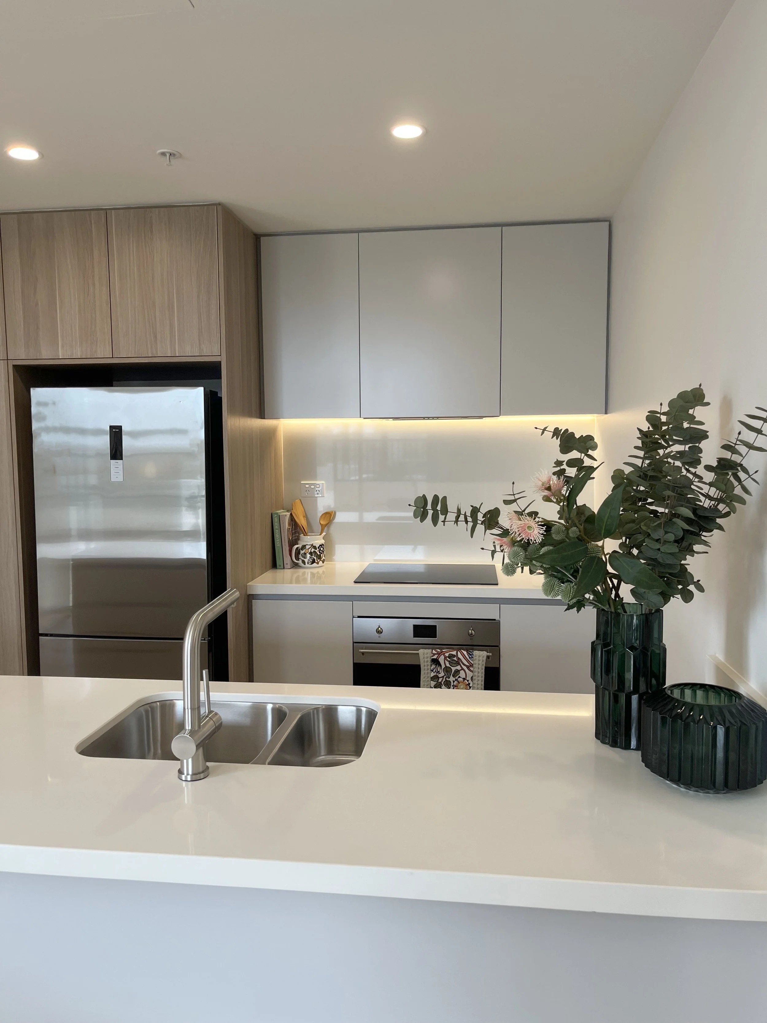 Modern kitchen with white countertop, stainless steel sink, green vases with flowers, wooden and white cabinets, and a stainless steel refrigerator.