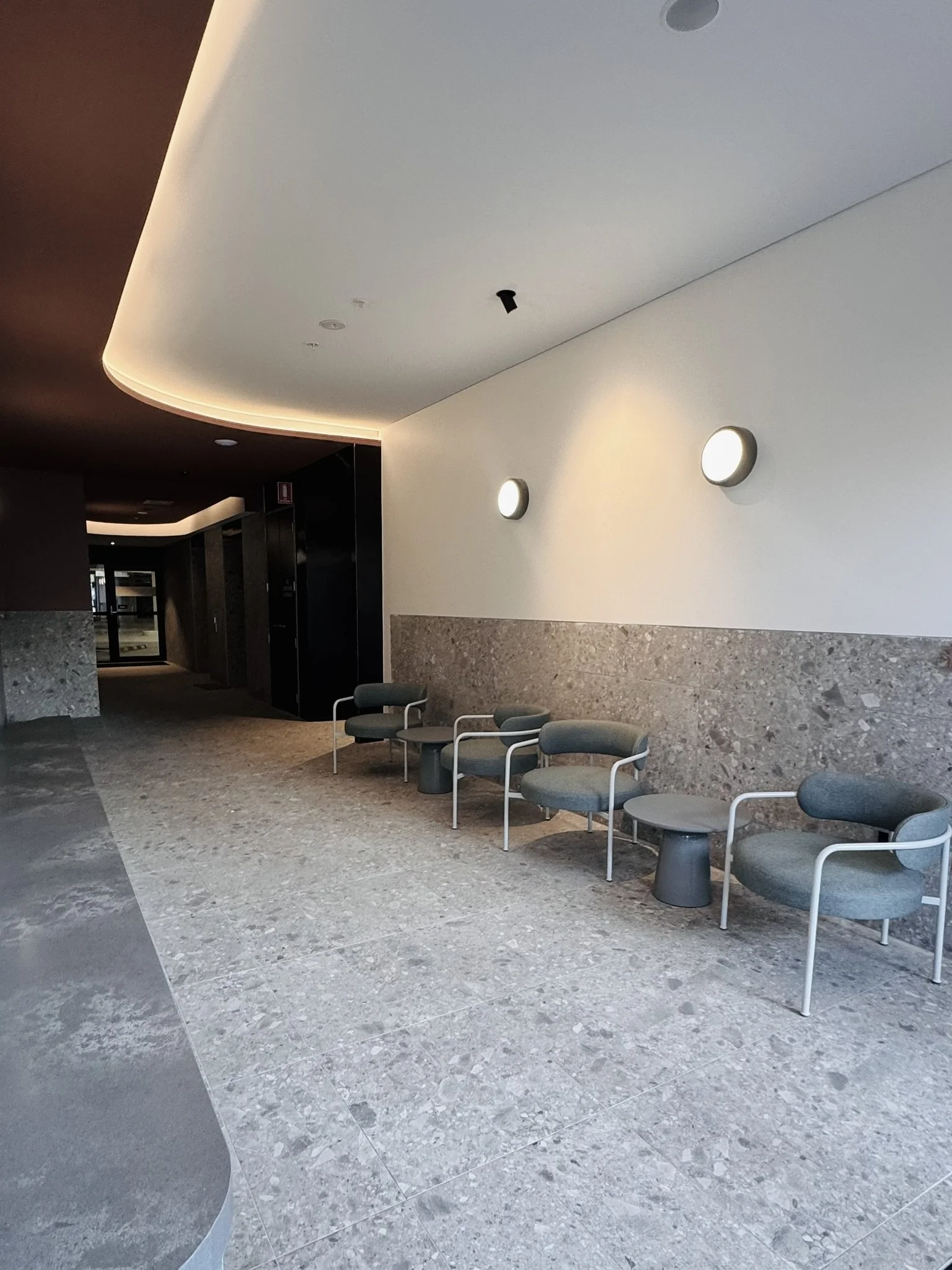 Empty modern lobby area with four gray chairs lined along a wall, small tables between chairs, marble flooring, white wall with circular wall lights, and a curved ceiling with soft lighting.