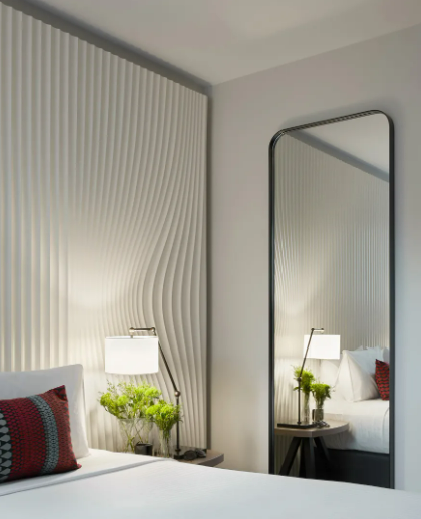 Interior of a modern bedroom with a bed, two lamps, a green plant on a nightstand, a large mirror, and textured white walls.