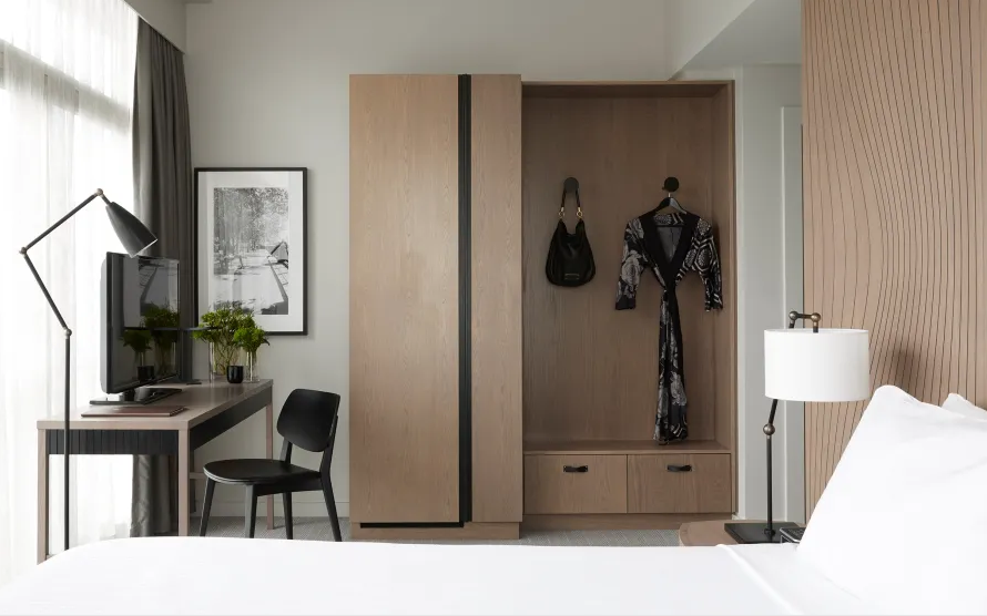 Interior of a bedroom with a wardrobe, a bedside lamp, a bed, a wall-mounted TV, a small desk with a chair, plants, and artwork on the wall.